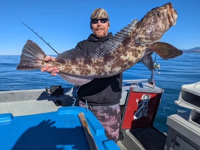Man holding a large fish on a boat in open water, wearing sunglasses, a camouflage hat, and a dark hoodie.