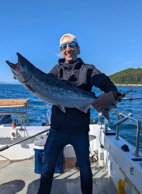 Man holding a large fish on a boat with water and trees in the background.