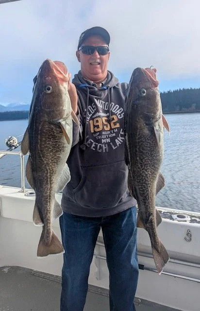 Man on boat holding two large fish, one in each hand, near a lake with mountains and trees in the background.