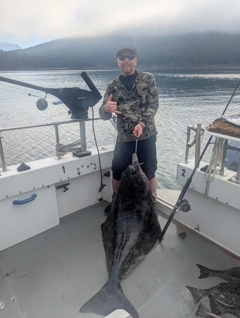 Man standing on a boat holding a large fish with a cloudy sky and mountains in the background.
