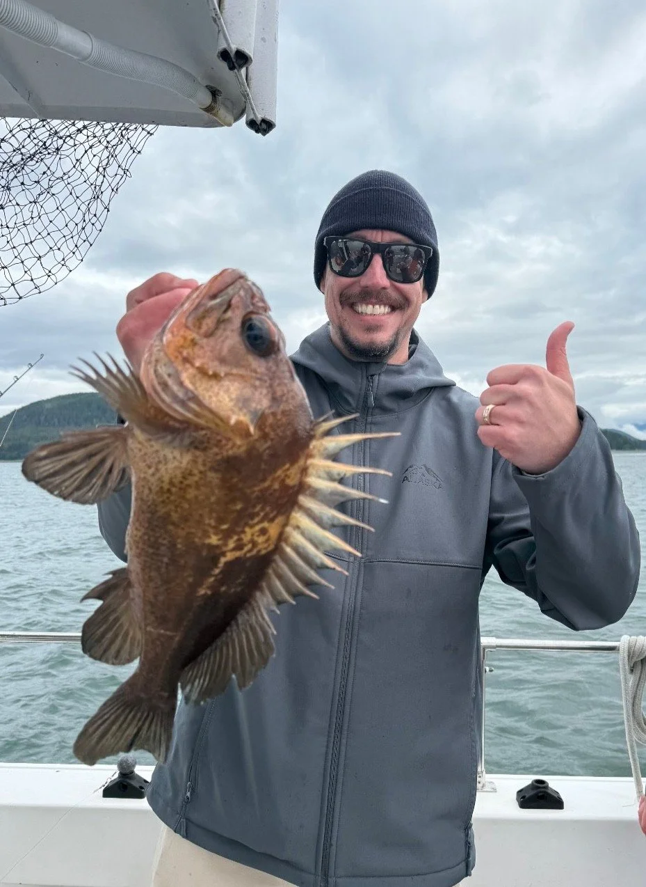 A man wearing sunglasses, a beanie, and a gray jacket on a boat holding a fish with spiny fins, showing a thumbs-up gesture with a cloudy sky and water in the background.