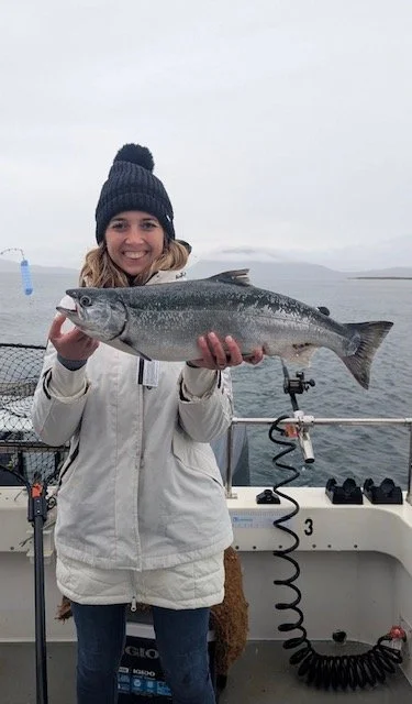 Woman in white jacket and black knit hat holding a large fish on a boat with water and cloudy sky in the background.