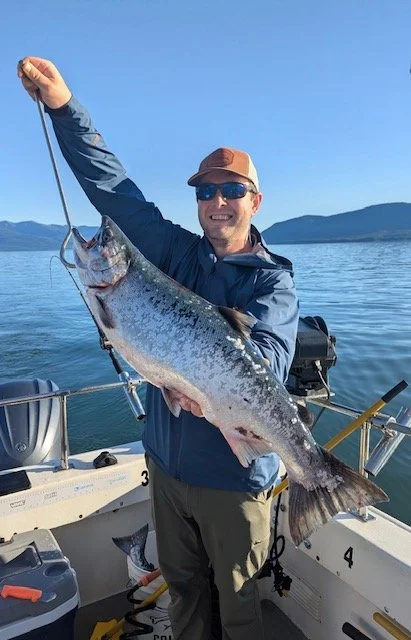A man wearing sunglasses, a brown cap, and a blue jacket holding a large fish he caught while on a boat in a lake, with mountains and a clear blue sky in the background.