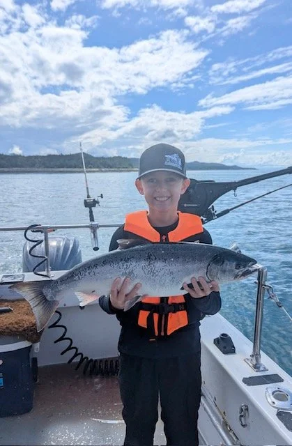 Young boy smiling on a boat holding a large fish he caught, with calm water, a boat motor, fishing rods, and a partly cloudy sky in the background.