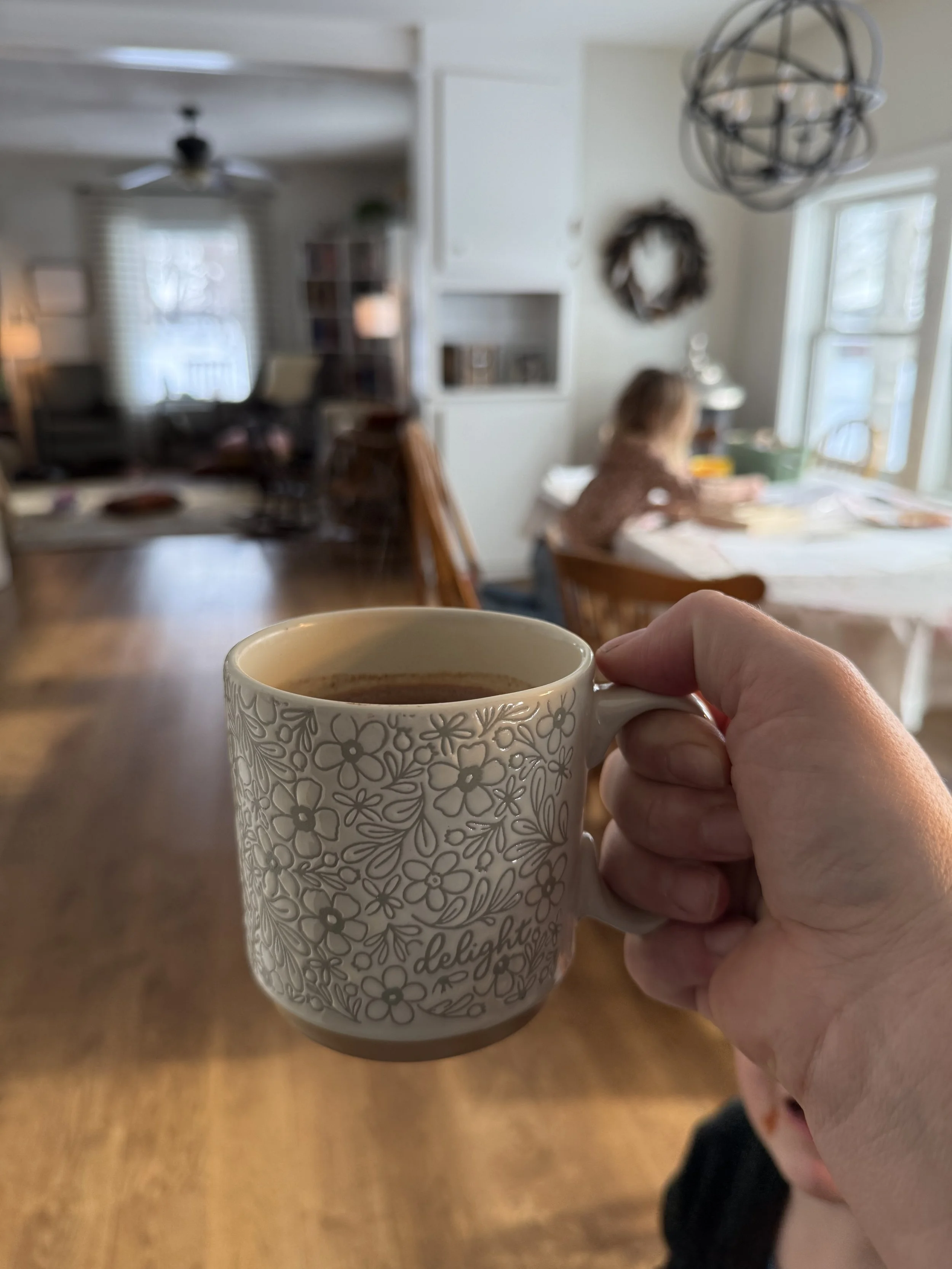 Person holding a mug with floral pattern and the word 'delight' while inside a cozy home with a child at a dining table in the background.