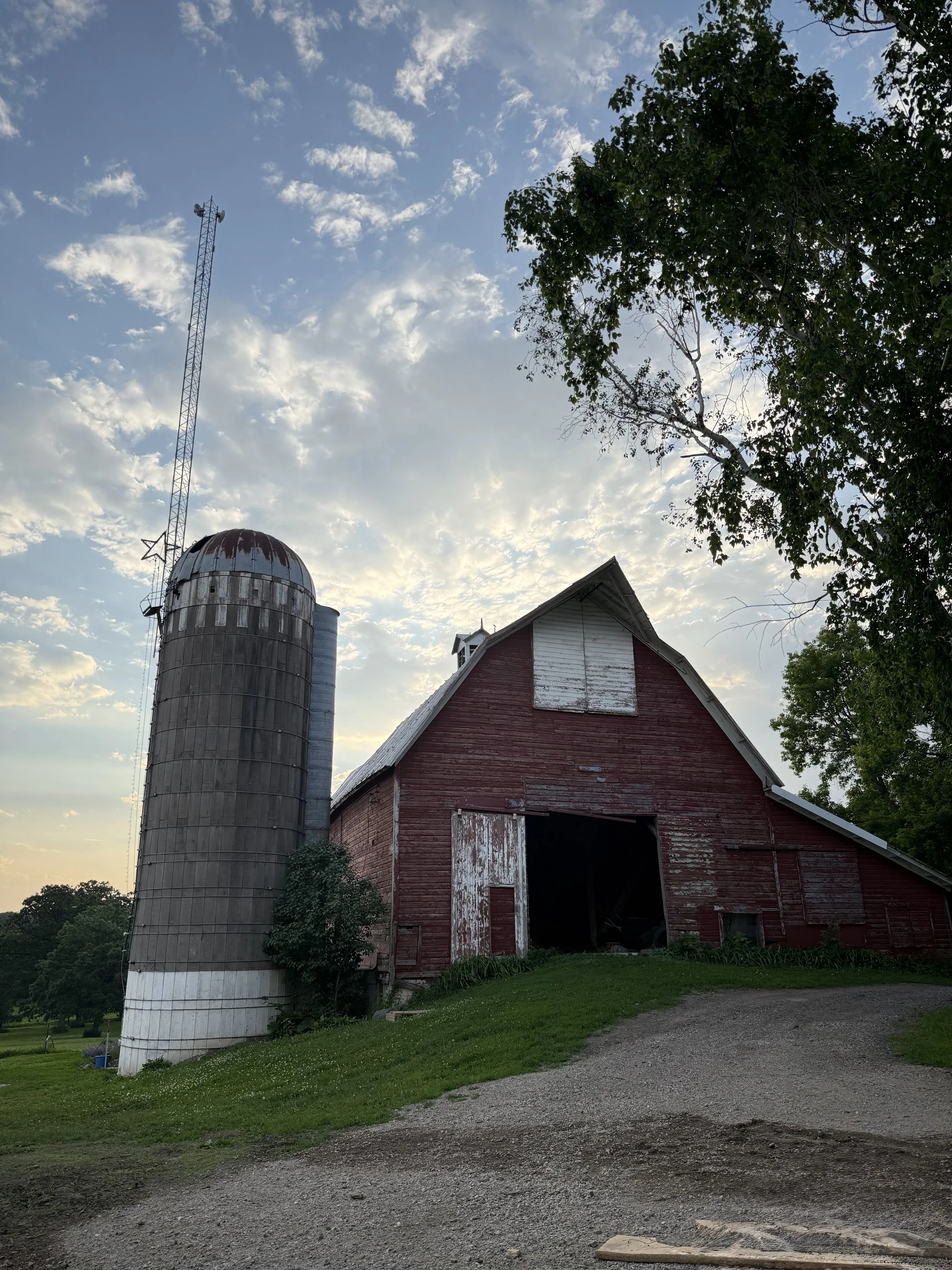 An old red barn with weathered, peeling paint next to a tall, rusty silo under a partly cloudy sky, surrounded by trees and green grass.