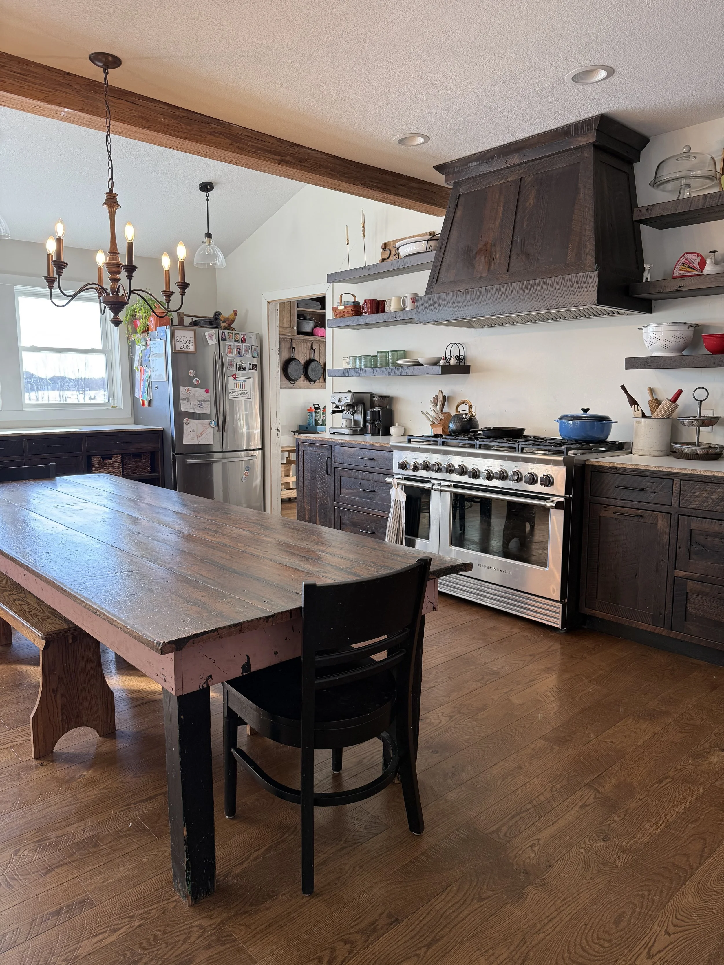 A rustic kitchen with a large wooden dining table, black chairs, dark wood cabinets, and a stainless steel oven. The room has open shelves with dishes and kitchenware, a coffee station, and hanging light fixtures. There is a window letting in natural