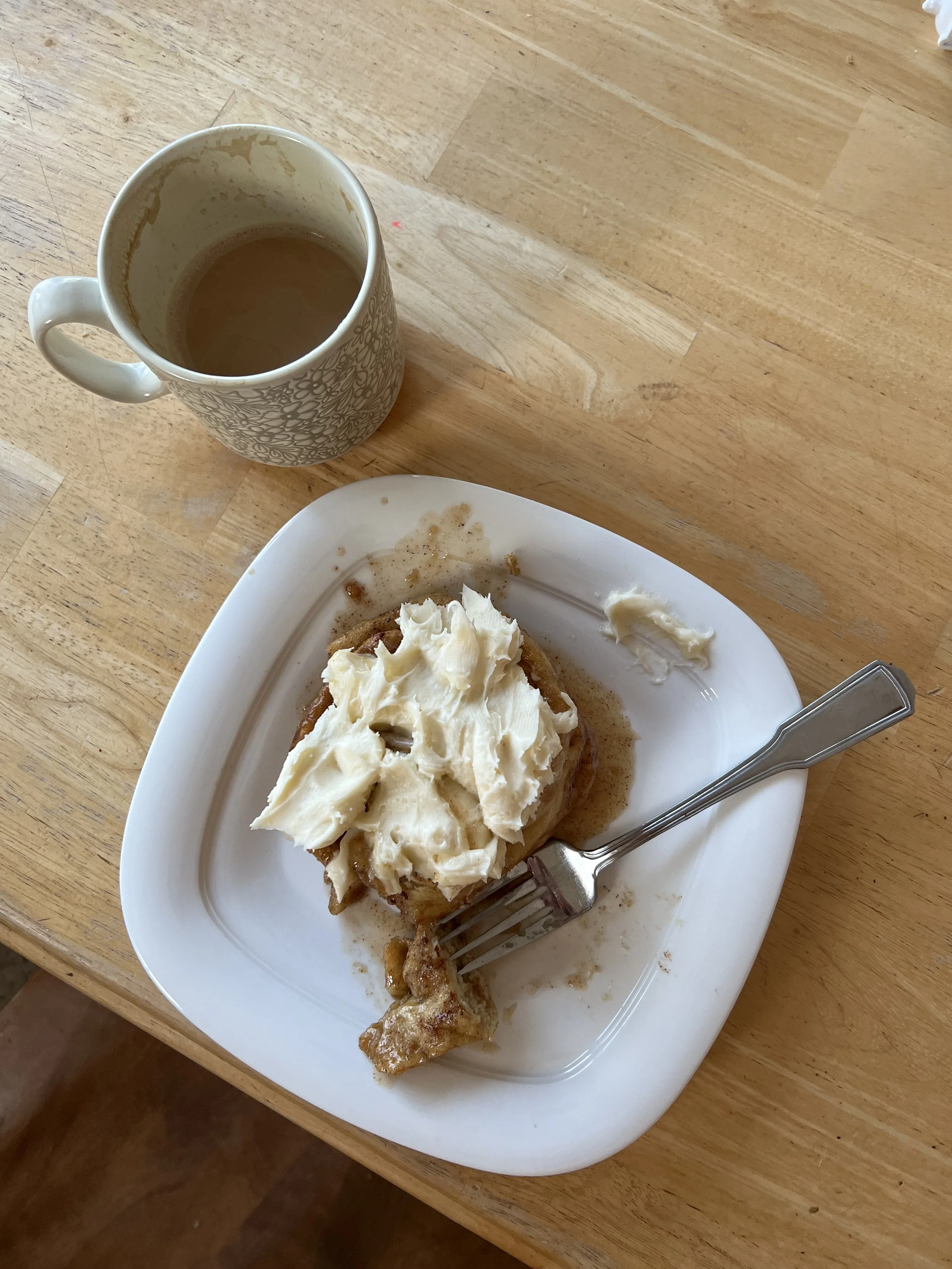 A half-eaten cinnamon roll with icing on a white plate, with a fork resting on it, and a mug of coffee on a wooden table.