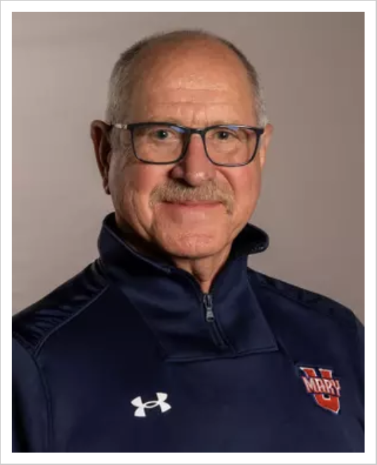 Portrait of an older man with glasses, gray hair, wearing a navy blue Under Armour jacket with a red, white, and blue team logo, smiling against a plain background.