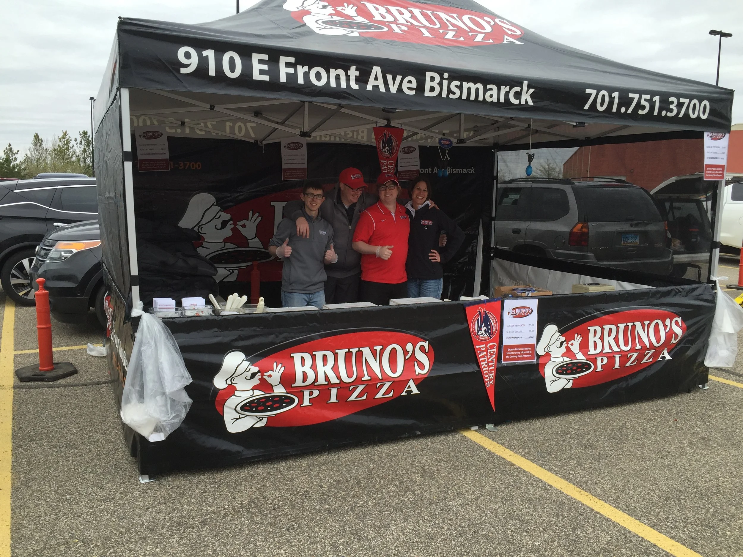 A group of four people standing inside a Bruno's Pizza tent in a parking lot. They are smiling, with two giving thumbs-up signs. The tent has black and red branding with the restaurant's logo and contact information.