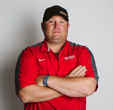 Man in a red sports shirt and black cap with arms crossed, standing against a white background.
