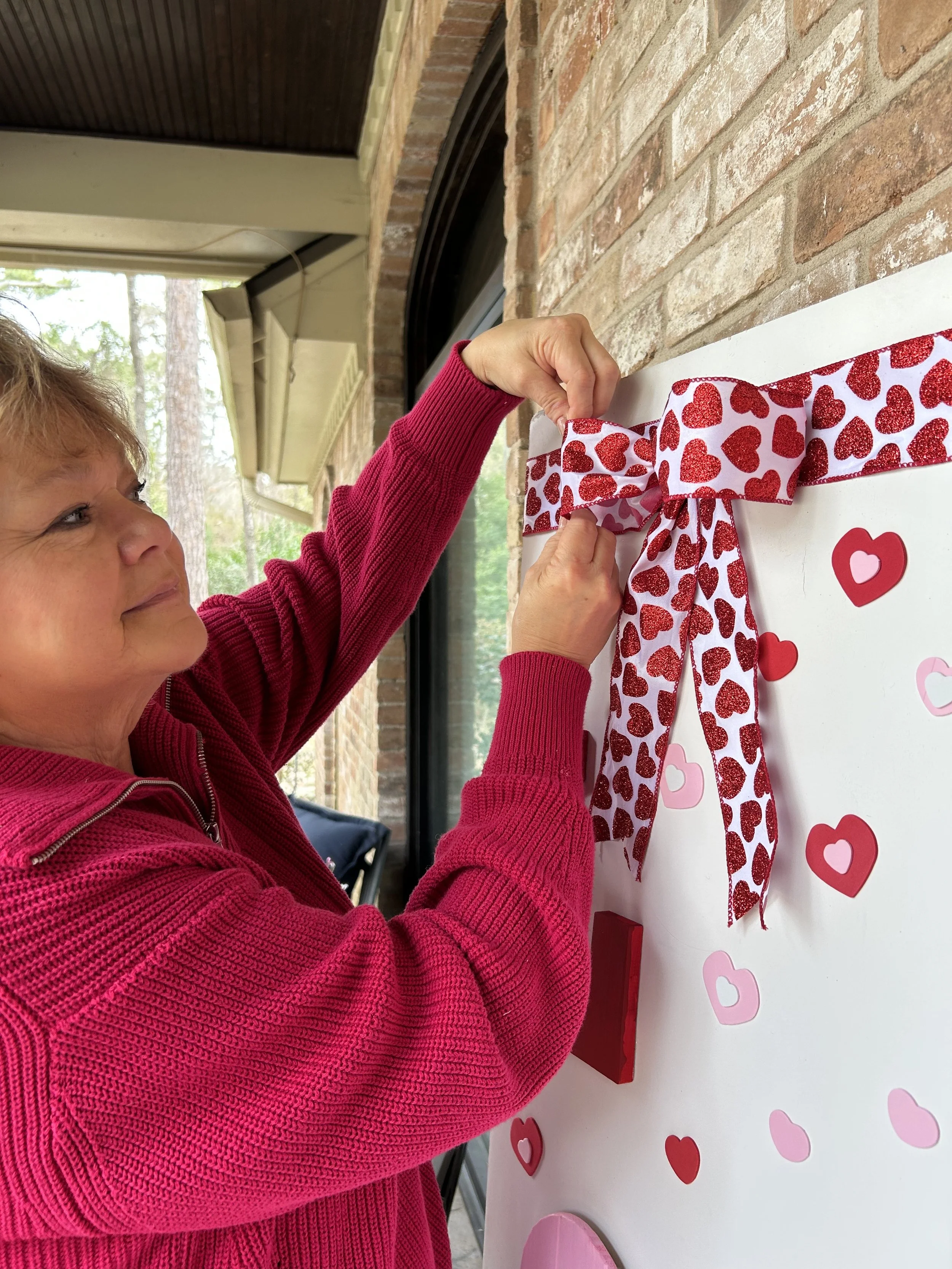 A woman in a red sweater is hanging a decorative bow with red glitter hearts on a white board decorated with heart-shaped stickers.