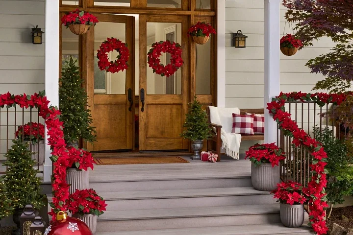 Front porch decorated for Christmas with wreaths on the doors, poinsettias in pots, and garlands on the railing, with two small Christmas trees and a bench with a blanket and pillows.