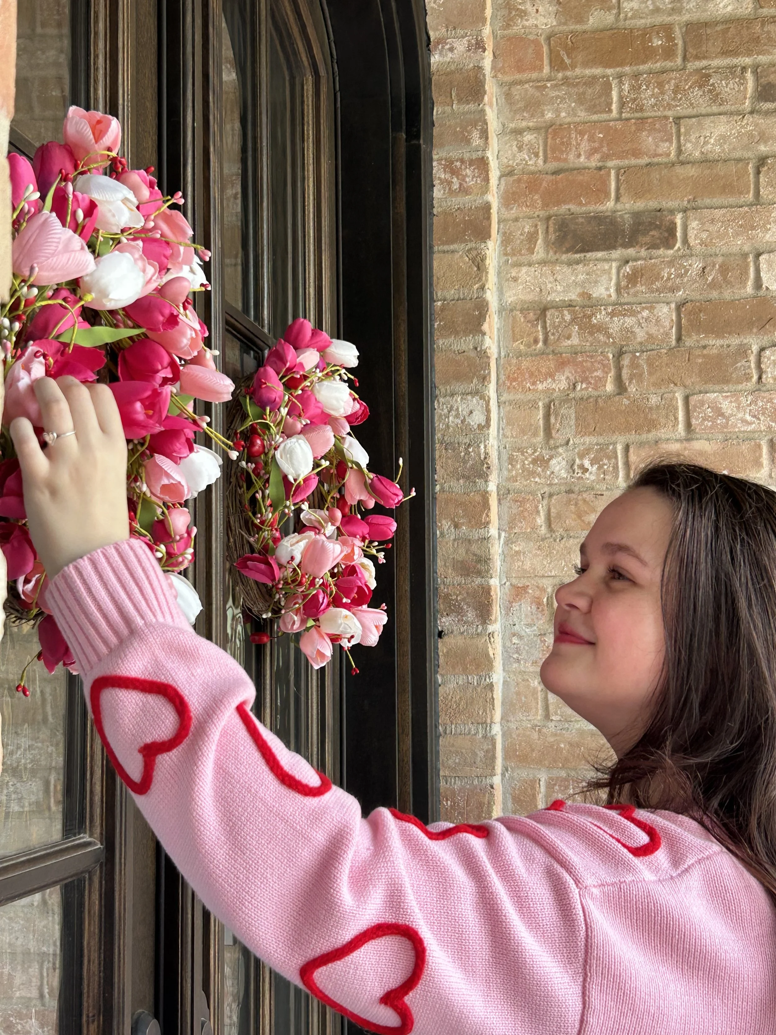 A girl in a pink sweater with red heart patterns on the sleeve is smiling and hanging a pink and white flower garland on a black metal gate in front of a brick wall.