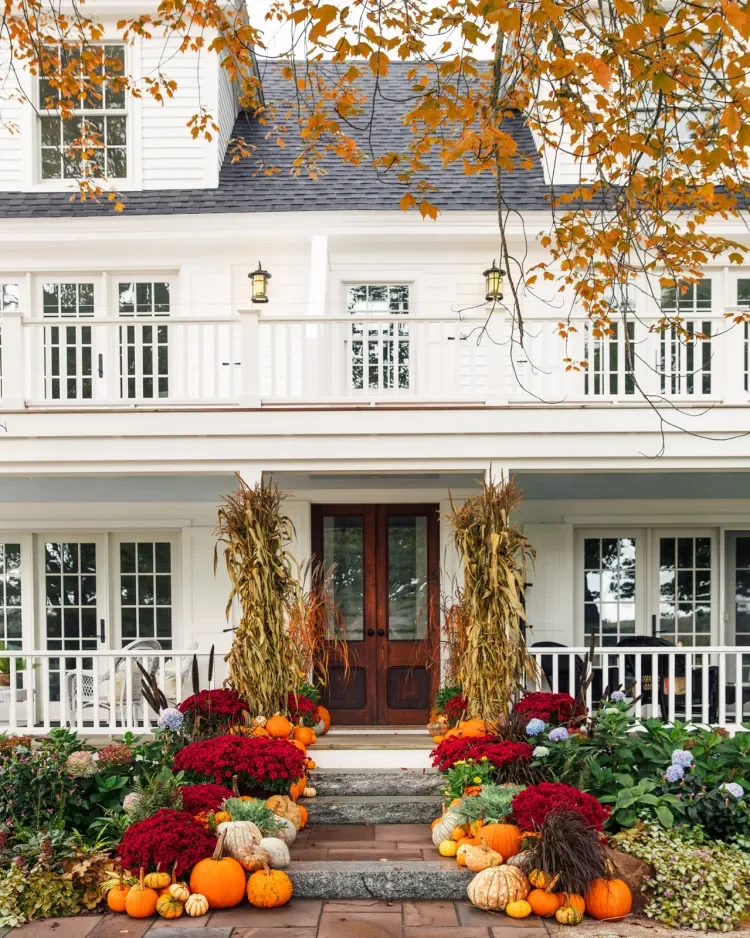 Front porch of a white house decorated for fall with pumpkins, gourds, and colorful fall flowers, framed by orange autumn leaves.