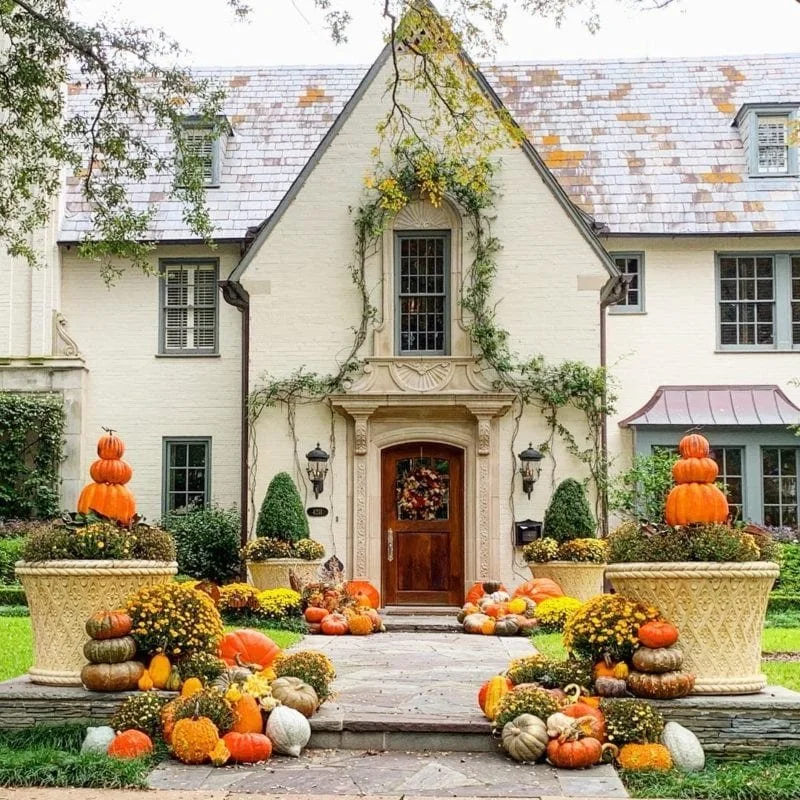 Front entrance of a house decorated with autumn harvest items including pumpkins, gourds, and chrysanthemums, with a pumpkin stack on each side of the pathway, and a door with a wreath.