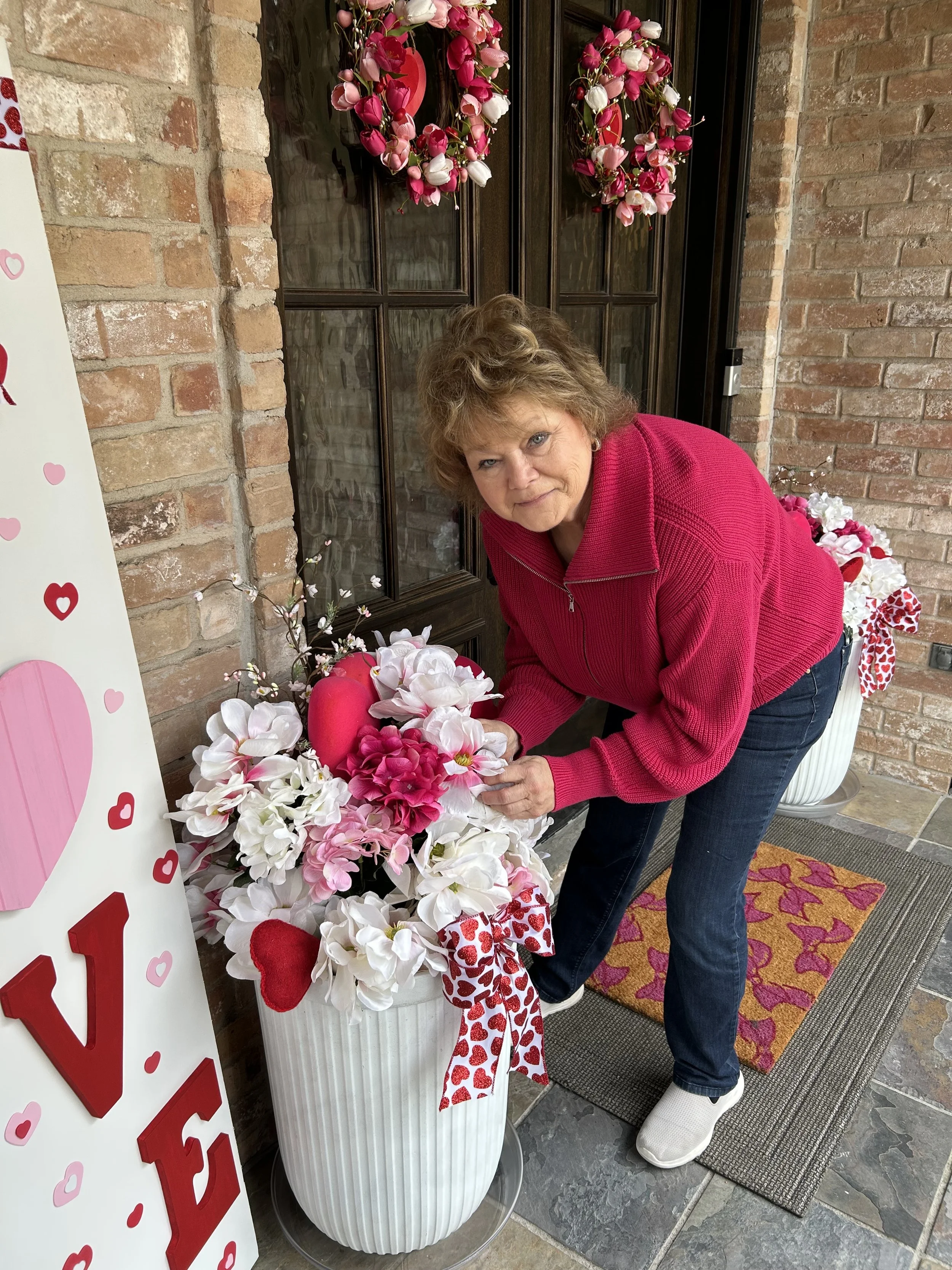 A woman in a pink jacket and jeans arranging pink and white flower arrangements with hearts and Valentine's Day decorations outside a brick building.