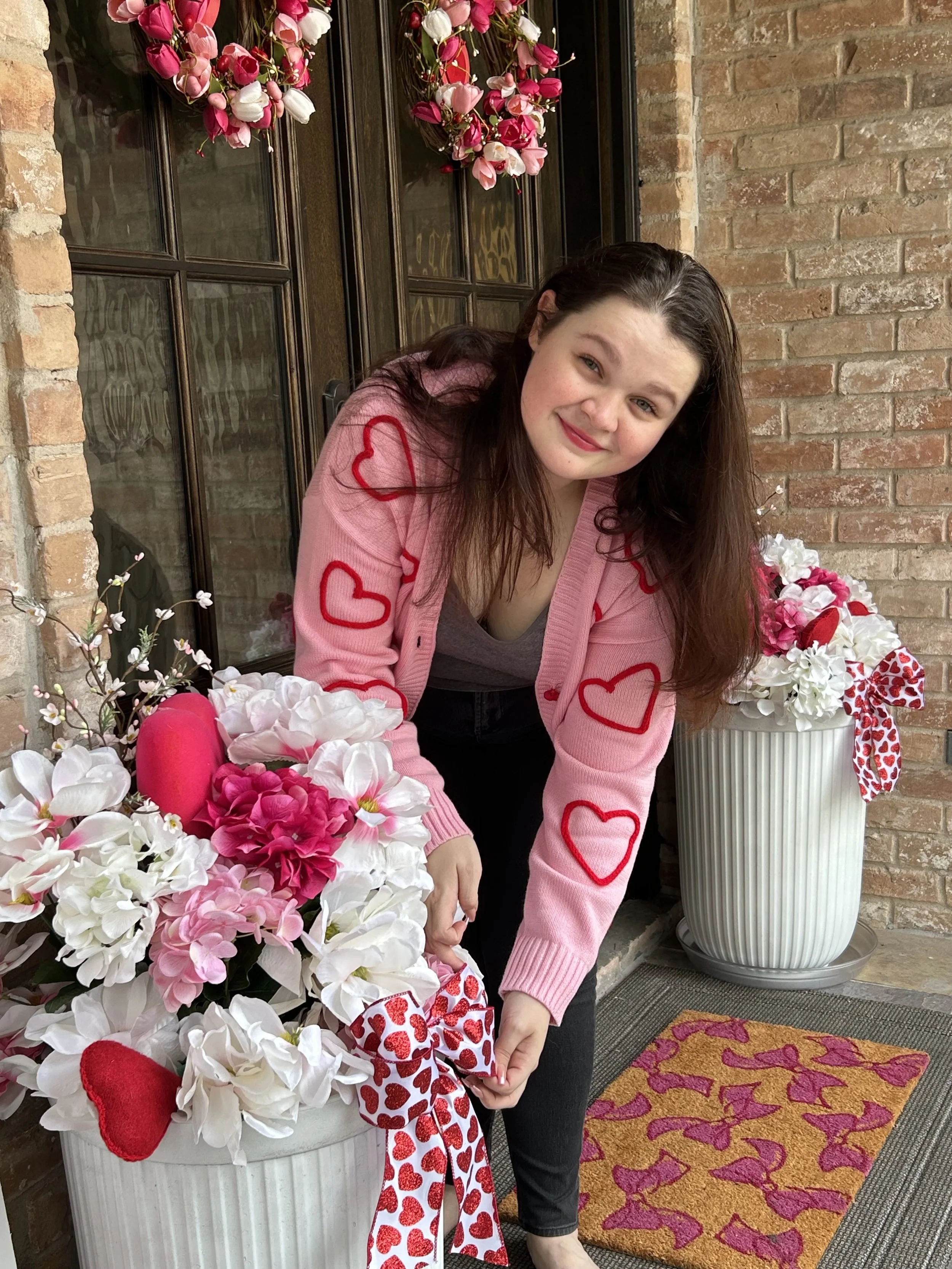 A woman with long brown hair and fair skin, wearing a pink cardigan with red hearts, bending down to adjust a white pot filled with pink and white flowers, on a porch with a brick wall, a door with hearts, and a Valentine's Day-themed doormat.