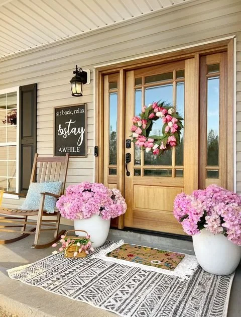 Front porch with a wooden door decorated with a pink flower wreath, two large pink flower pots, a rocking chair, a chalkboard sign, and a colorful mat.
