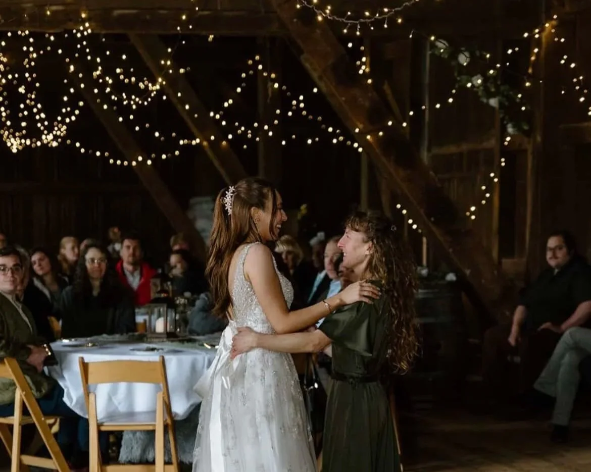 A bride and a woman dancing and smiling at a wedding reception with string lights overhead and guests seated at tables in the background.