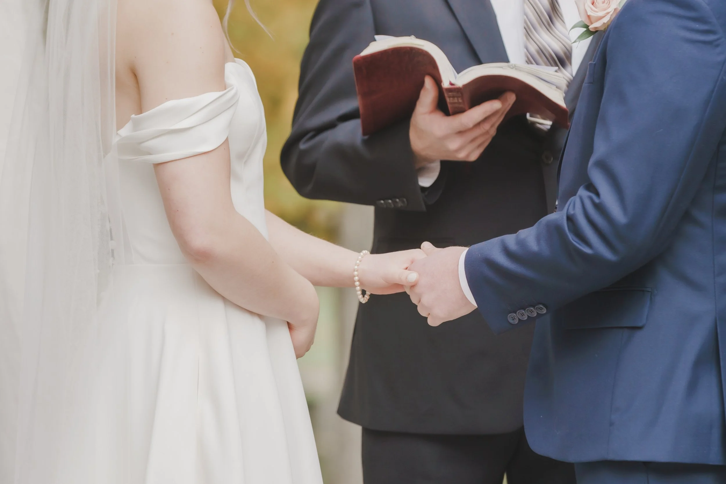 A bride and groom holding hands during their wedding ceremony, with an officiant reading from a book.