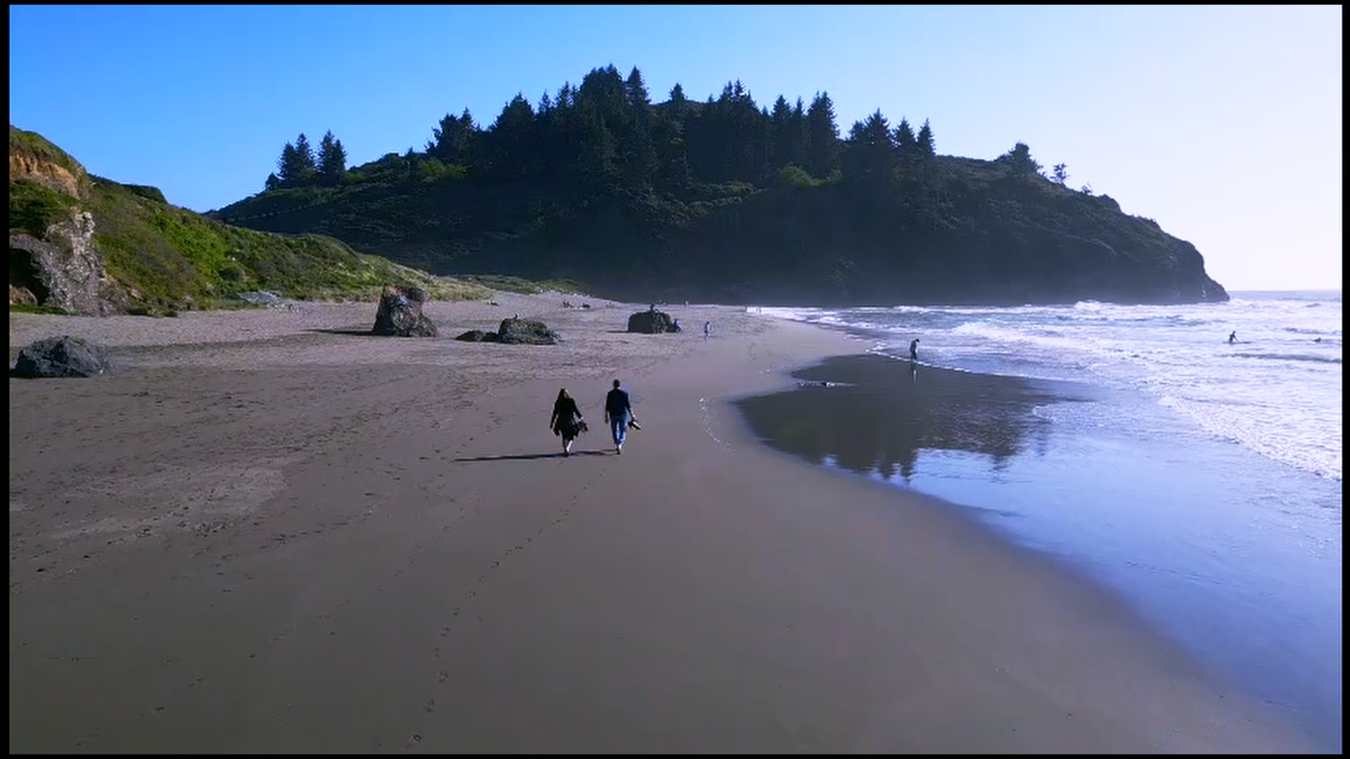 Couple walking on a sandy beach near the ocean with rocks and green forested hills in the background, under a clear blue sky.