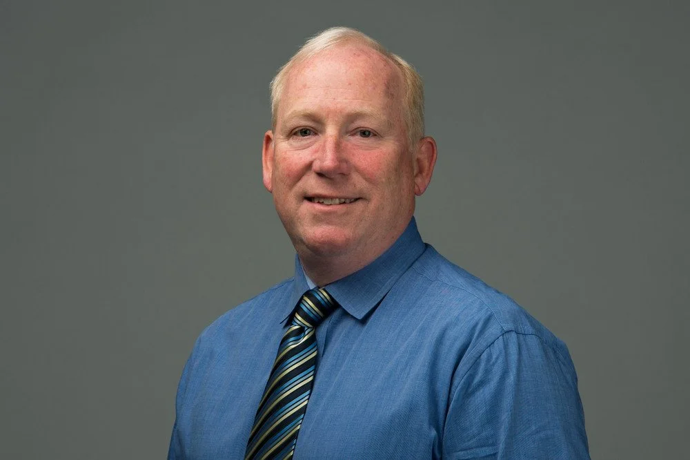 Portrait of a middle-aged man with short light hair, wearing a blue dress shirt and a striped tie, standing against a plain gray background.