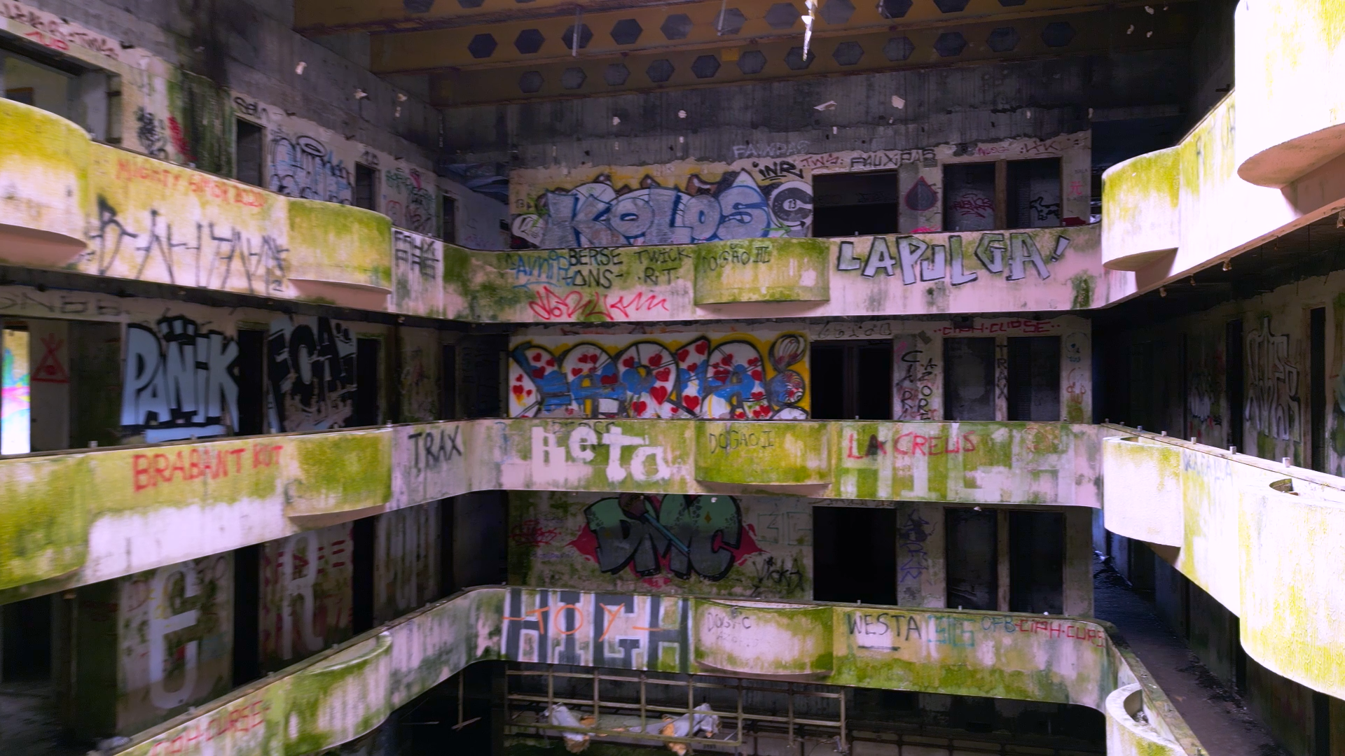 Abandoned, multi-story building with graffiti-covered walls, peeling paint, moss, and broken windows.
