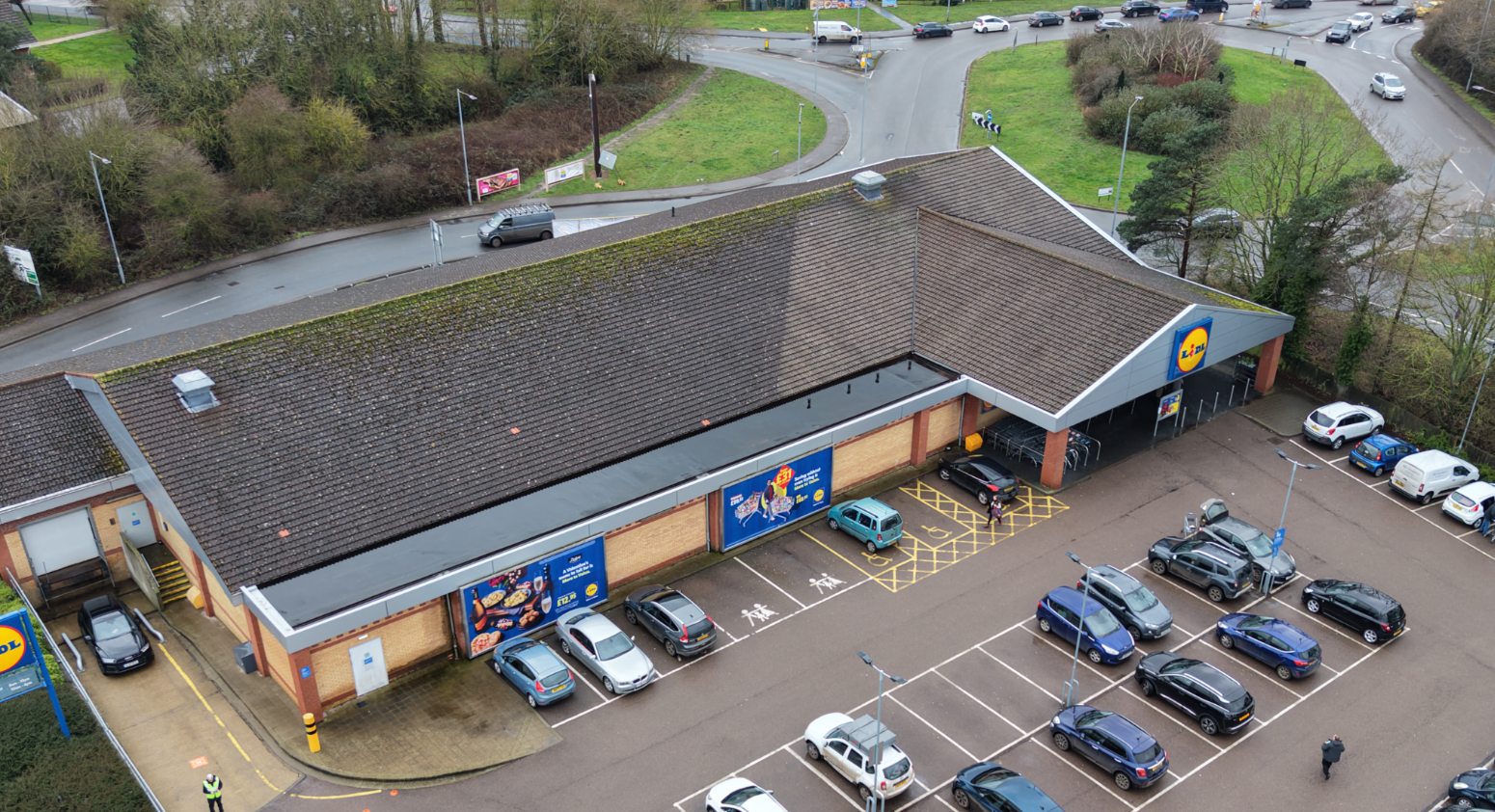 Aerial view of a Lidl supermarket with a parking lot in front, several cars parked, and a few people near the entrance. Surrounding roads and green areas are visible.