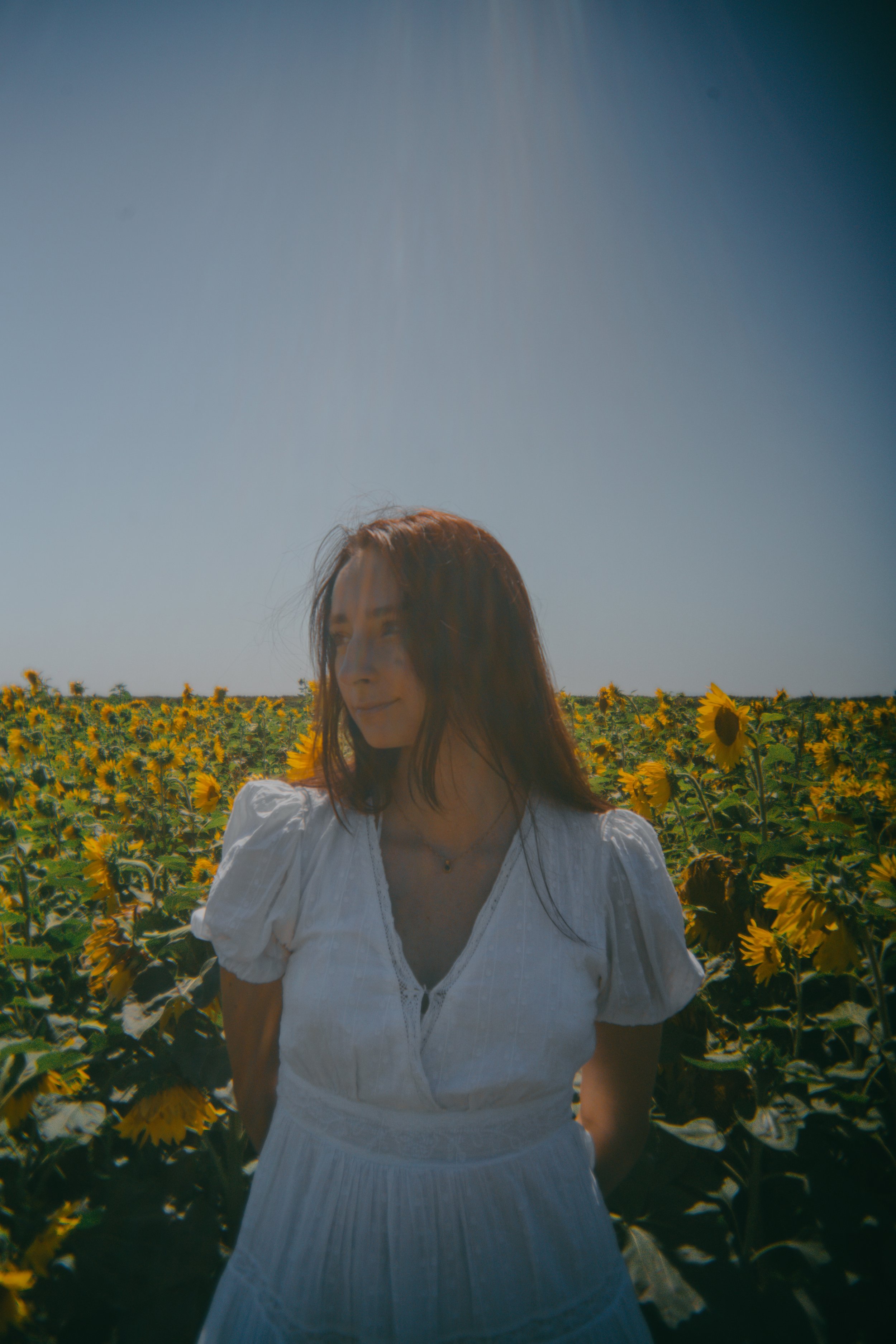 A woman with long hair stands in a sunflower field under a clear blue sky.