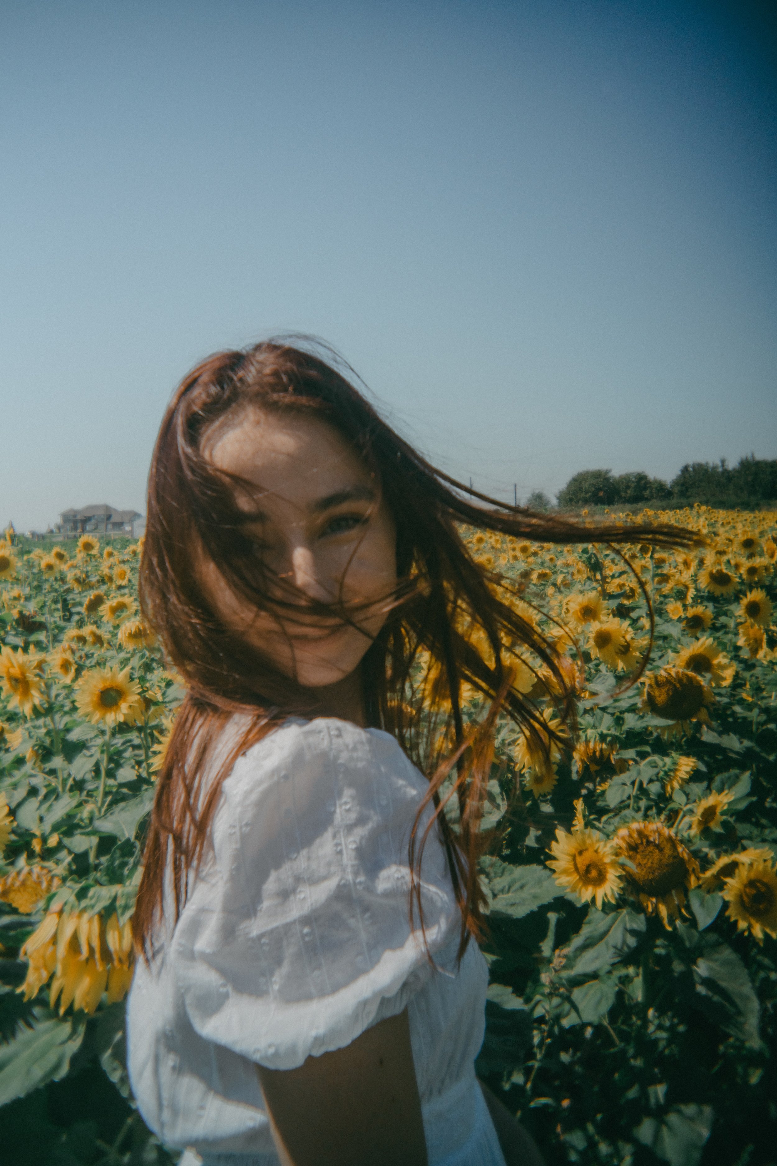 A woman with long red hair smiling in a sunflower field under a clear blue sky.