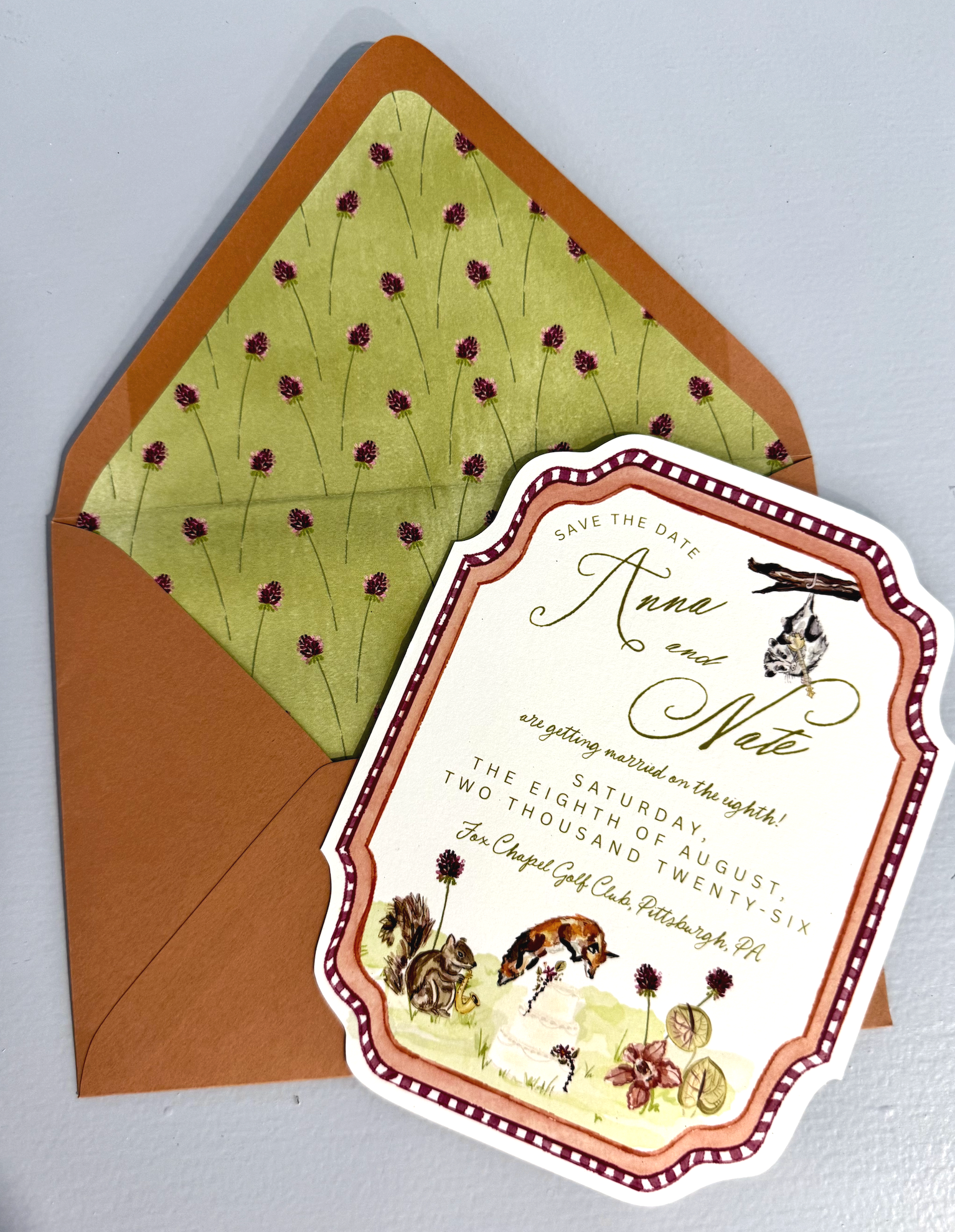 Wedding save-the-date card with floral and animal illustrations, featuring an orange and white border and a woodland theme, placed on a matching floral envelope.