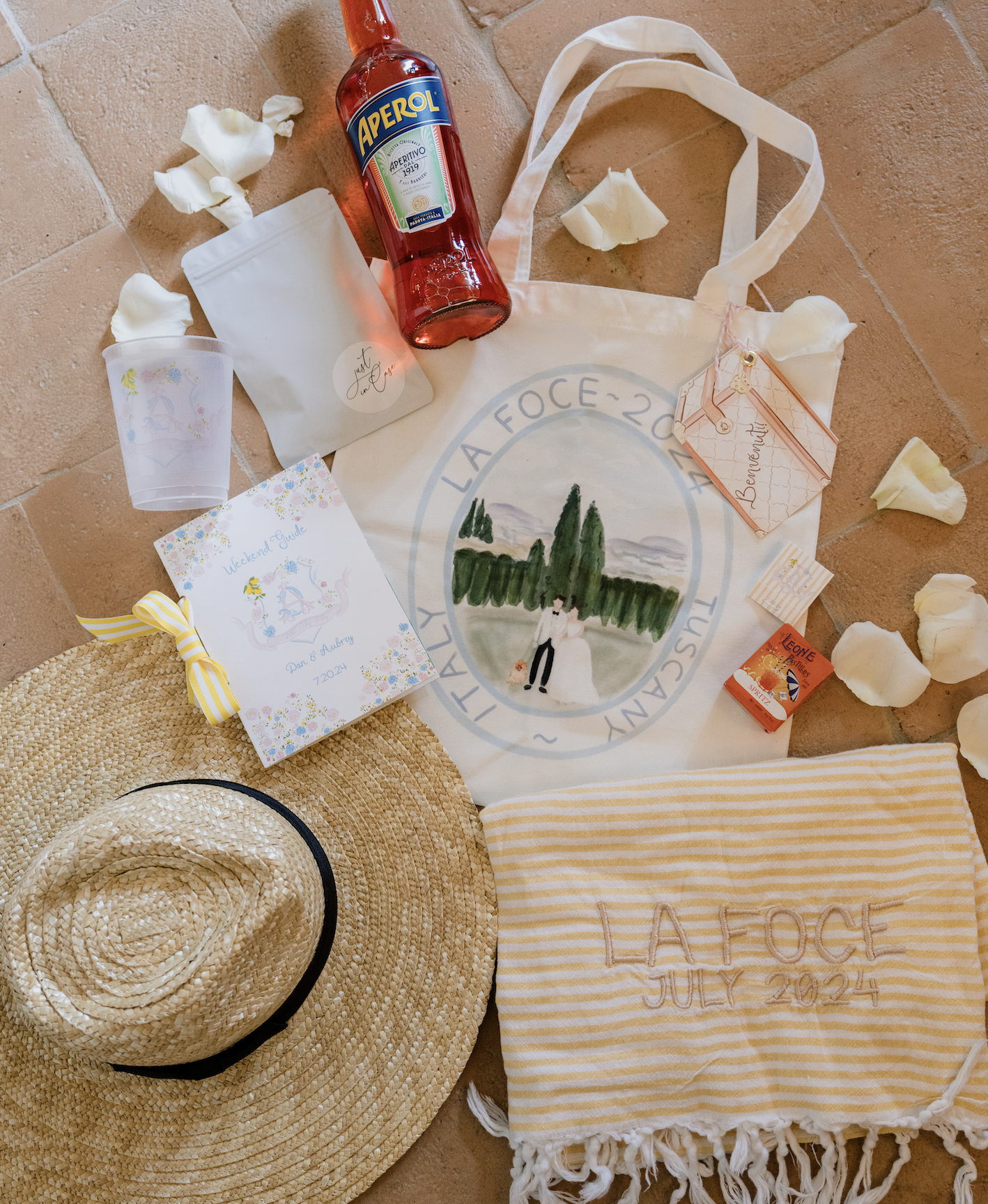 Summer wedding items including a straw hat, a striped towel with embroidered text, a tote bag with a painted scene and text, a bottle of Aperol, a small booklet, a plastic cup, a package, a box of Leone matches, and scattered flower petals.