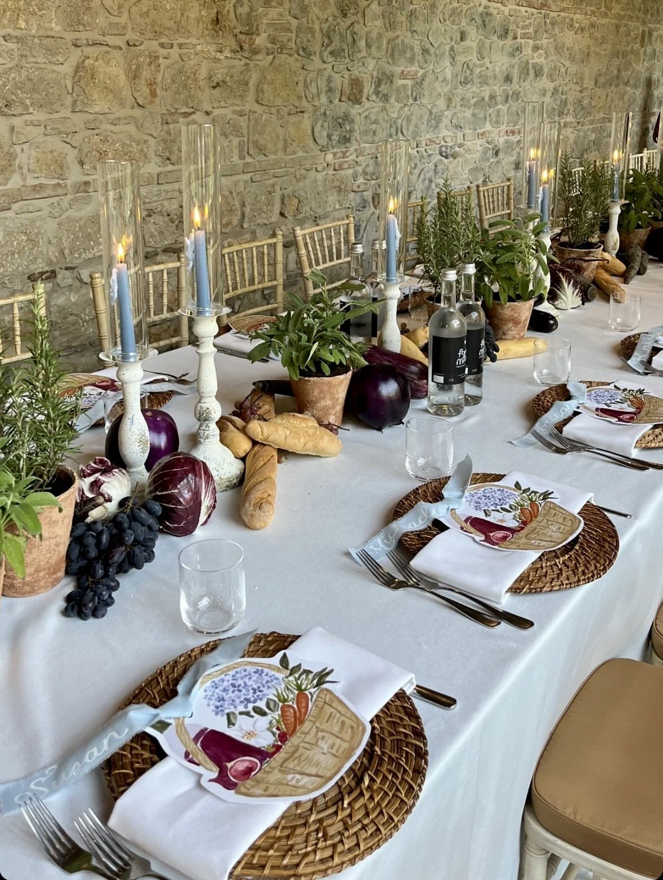 Table setting with woven placemats, white napkins, and decorative plates featuring vegetables, in front of a stone brick wall, decorated with candles, potted plants, bread, and bottles.