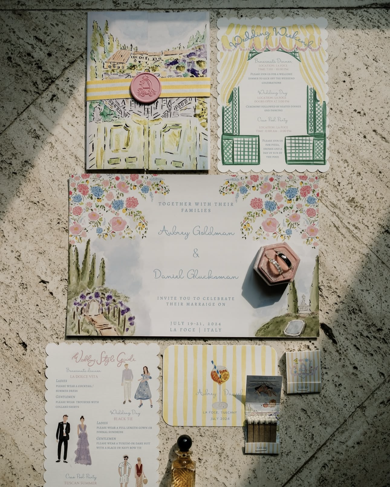 Wedding invitation suite with floral and scenic illustrations, rings in a pink velvet box, a miniature bottle of alcohol, and flowers on a textured surface.