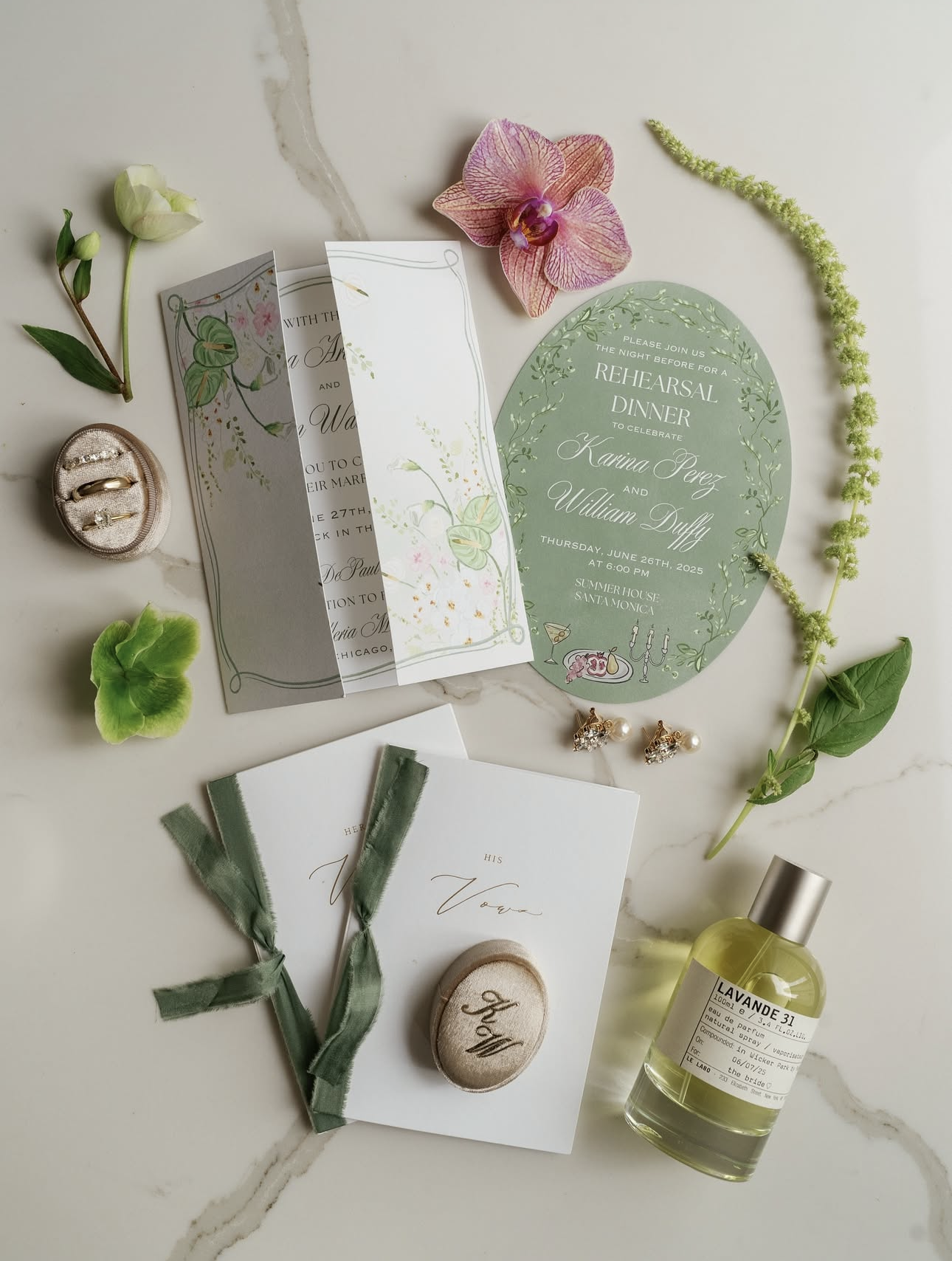 Wedding invitation suite with floral and botanical accents, jewelry, green ribbons, perfume bottle, and decorative flowers arranged on marble surface.