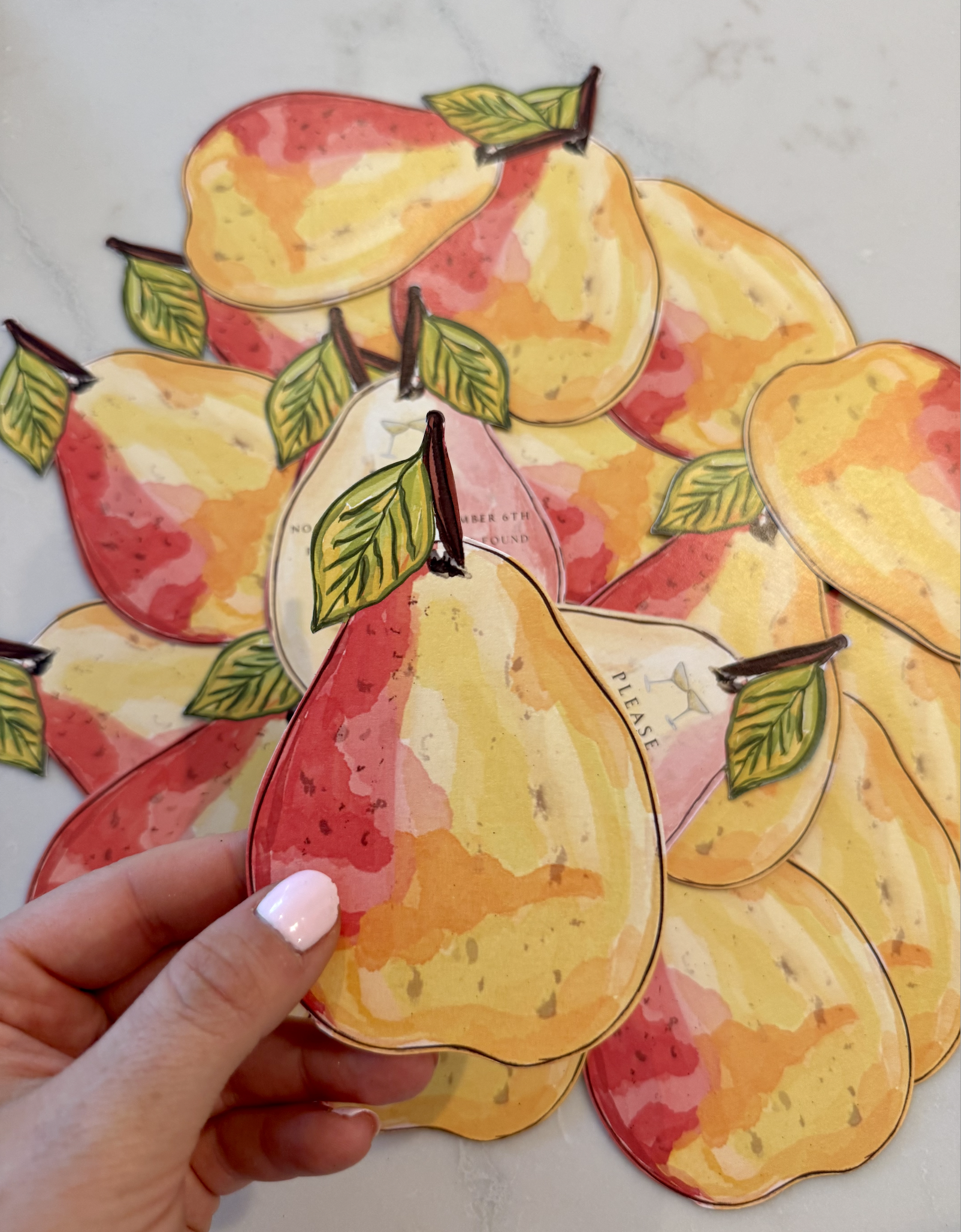 A person holding a paper cutout of a peach with green leaves, with several similar peach cutouts scattered on a white surface.