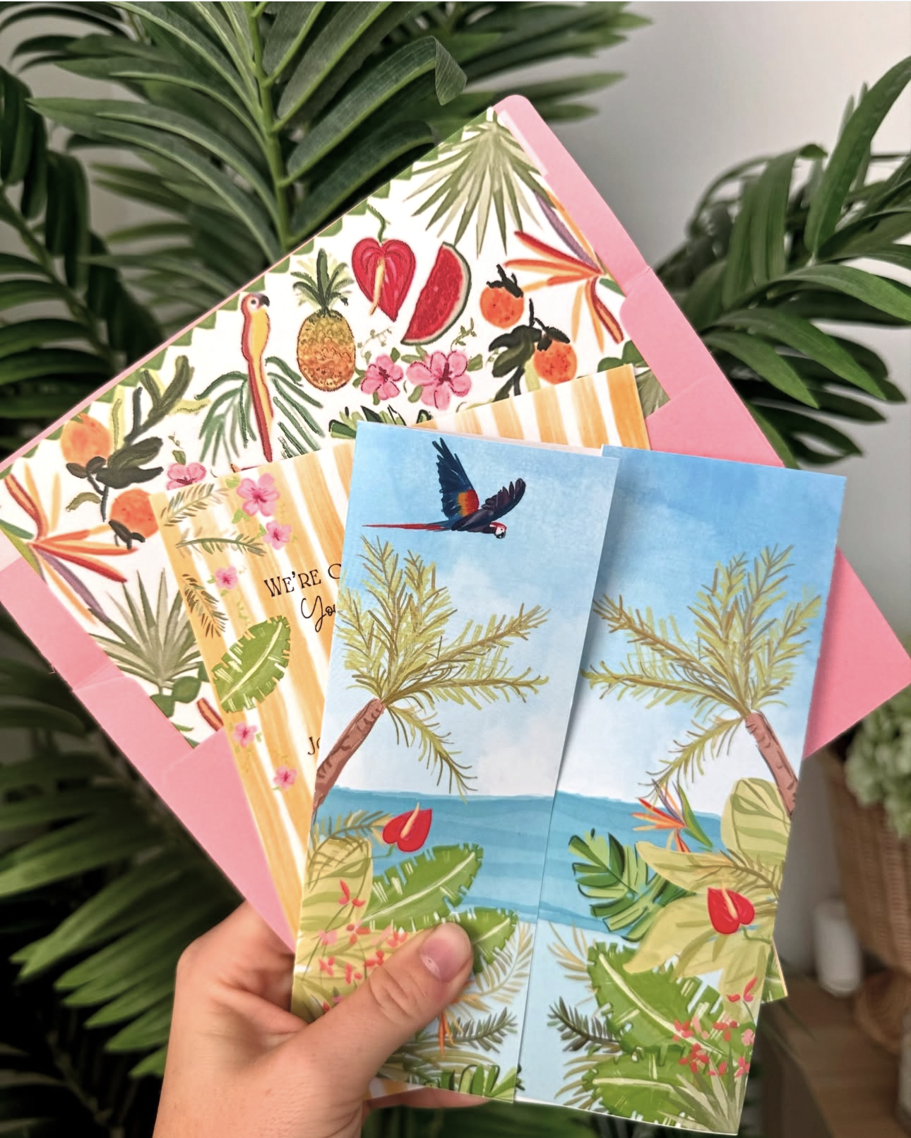 A hand holding tropical-themed greeting cards and a pink envelope with colorful fruit, plants, and birds, against a leafy green background.
