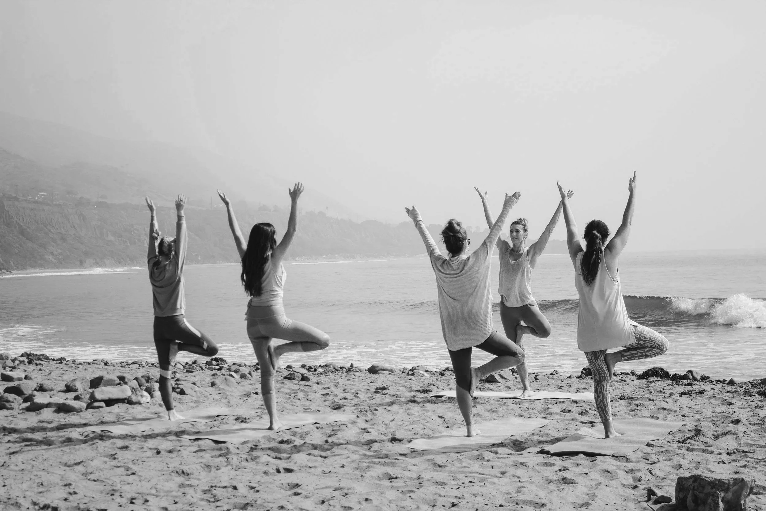 A group of five women practicing yoga on a beach, standing on mats, with arms raised and one leg bent at the knee, facing the ocean.