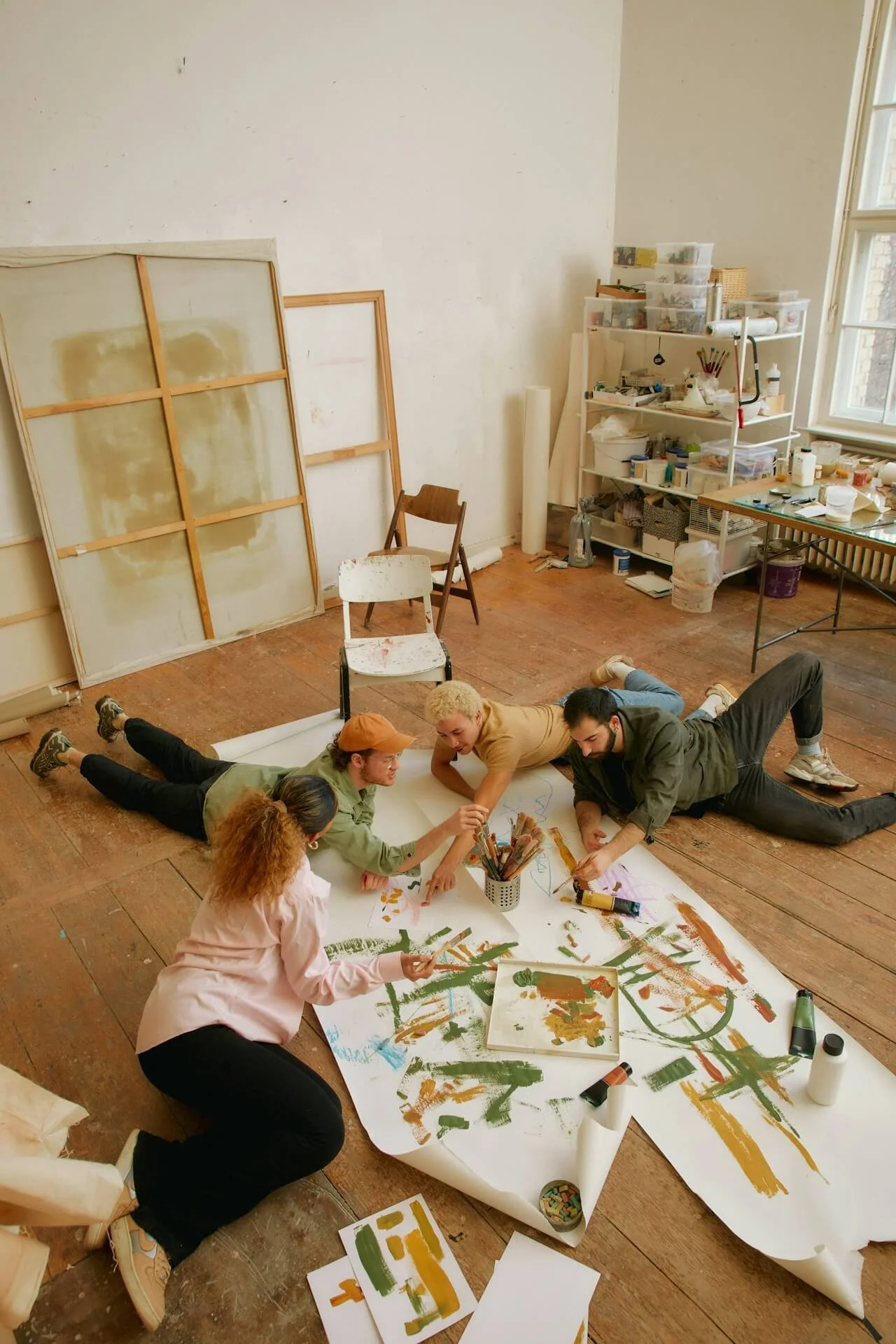 Five people laying on the wooden floor of an art studio, working together on a large abstract painting on a wide sheet of paper, surrounded by art supplies.