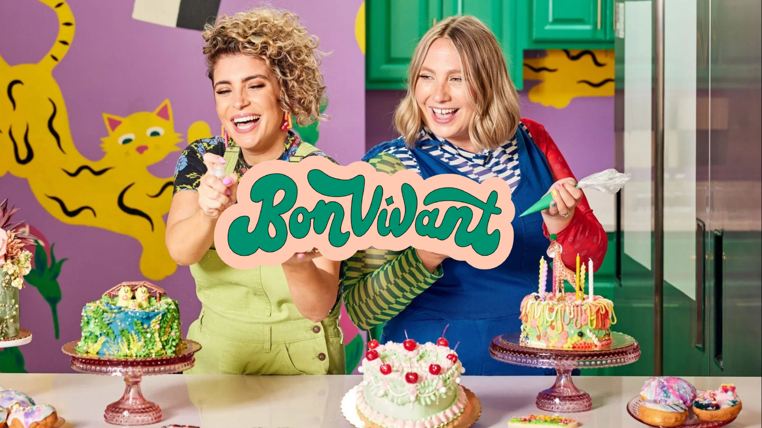 Two women at a birthday celebration with colorful decorated cakes and desserts on a table. One woman is holding a cake with a candle, and both are smiling. The background features a vibrant purple wall with yellow tiger designs.