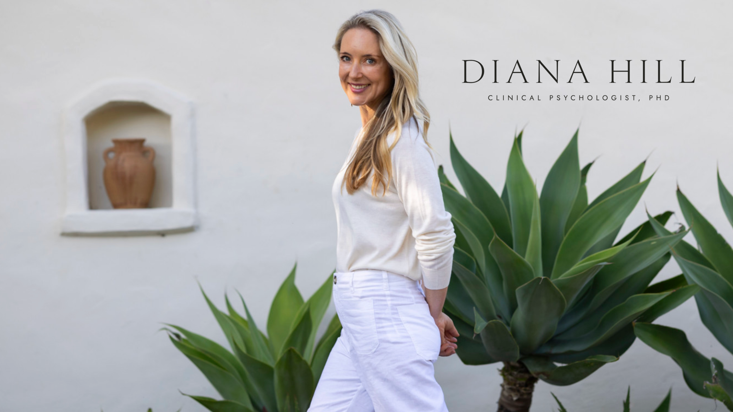 A woman with long blonde hair wearing a white long-sleeve shirt and white pants, standing next to large green succulent plants against a light-colored wall with a small recessed shelf containing a decorative vase.