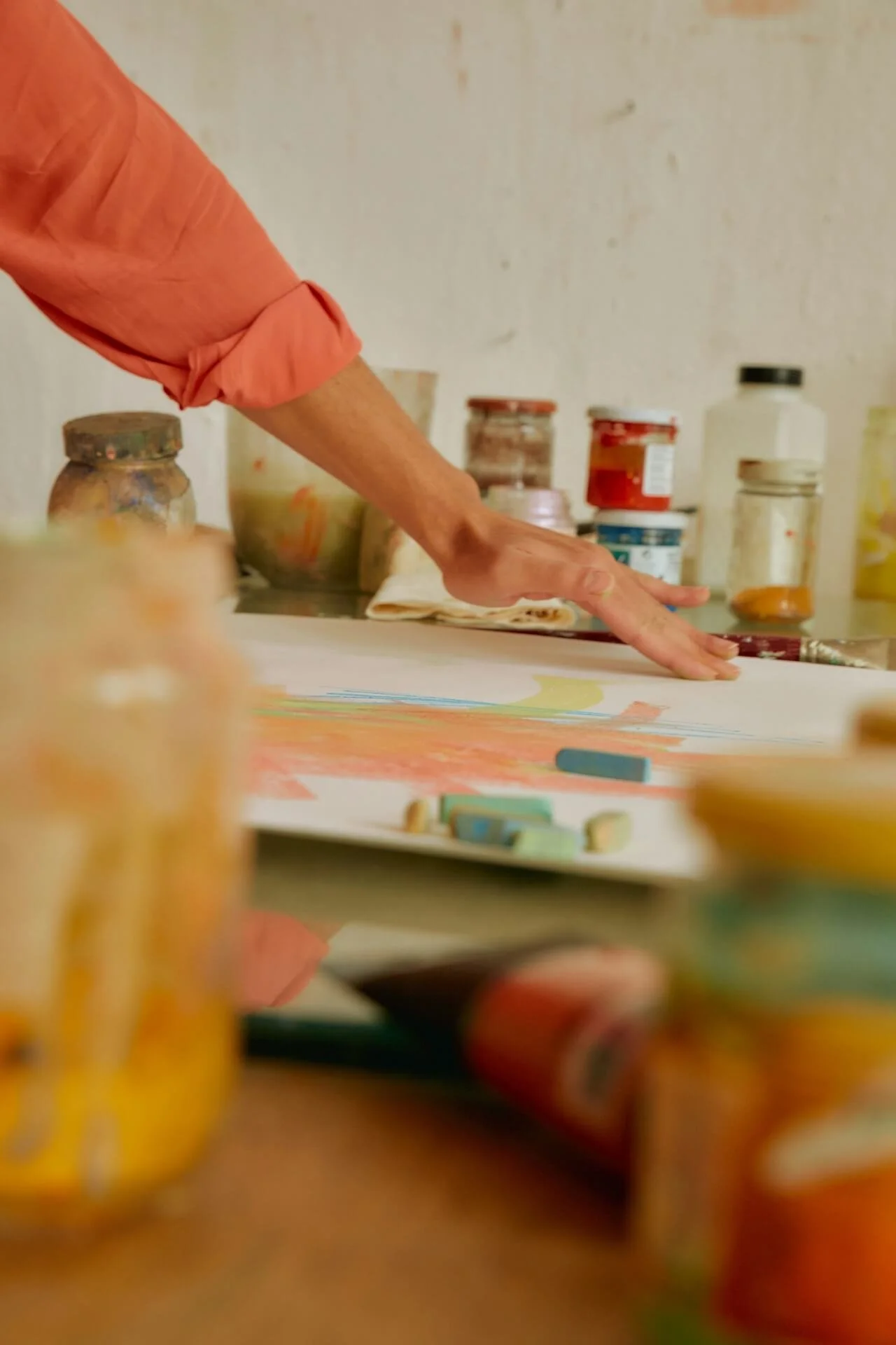 A person wearing a coral-colored long-sleeve shirt is creating art on paper with chalks. There are various jars of paint or art supplies on the table, and the person is reaching across the artwork on the table surrounded by colorful chalks and art materials.