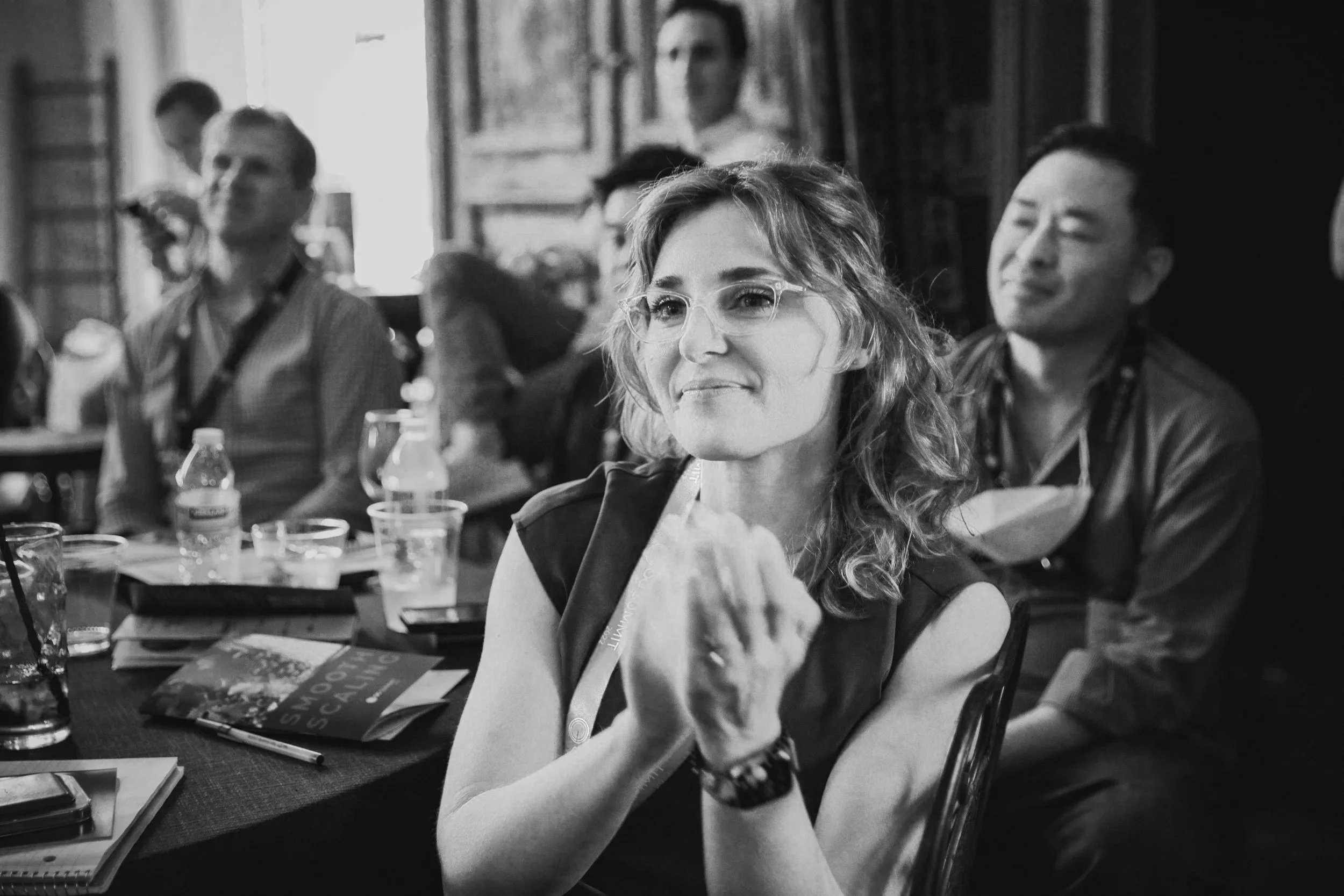 A woman with wavy hair and glasses sitting at a conference table, smiling slightly and holding her hand near her face. Other people are sitting around her, some with bottled drinks and notebooks, in a room with wood-paneled walls. 