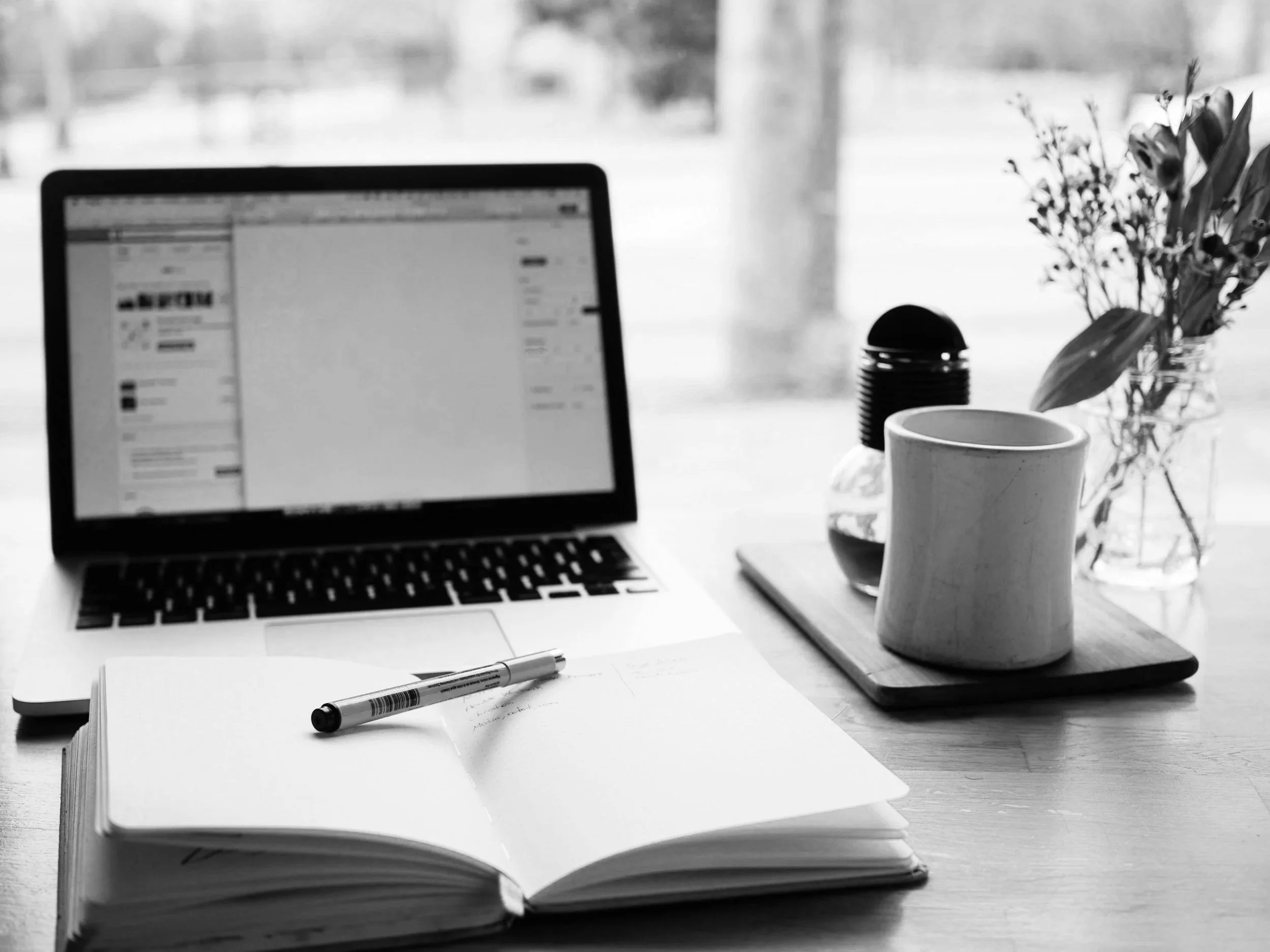 Black and white photo of a workspace with a laptop, open notebook with a pen, a coffee mug, a vase with flowers, and a small jar. The background is blurred. Course Creators can sell event tickets on Squarespace with Eventually.