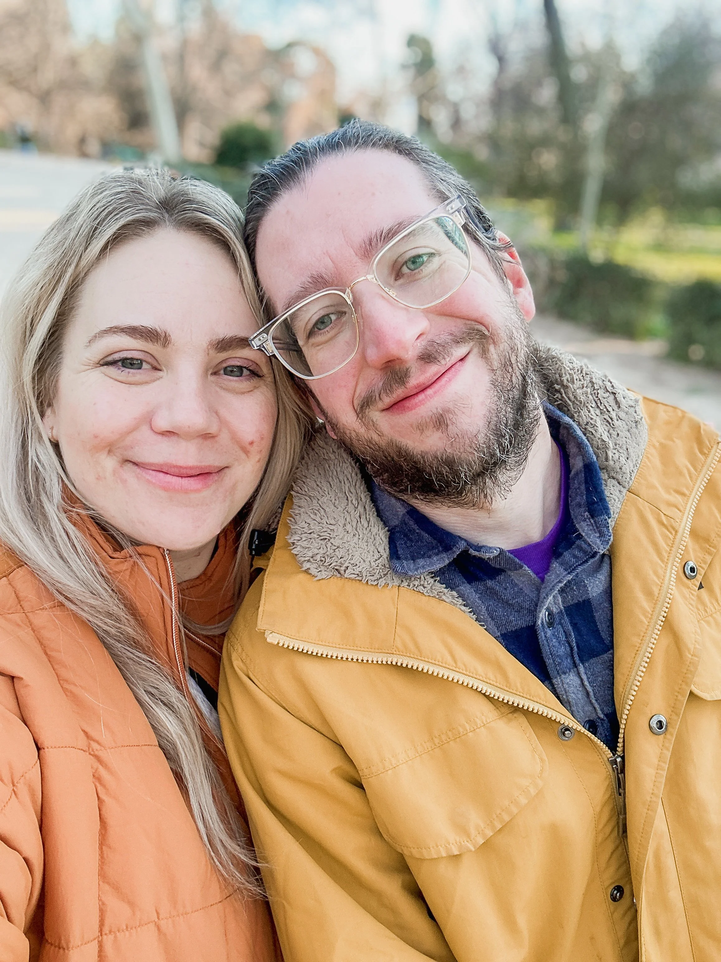 A close-up photo of two smiling people outdoors on a sunny day, with trees and a street in the background. The woman has blonde hair and is wearing an orange jacket, and the man has dark hair, glasses, a beard, and is wearing a yellow jacket.