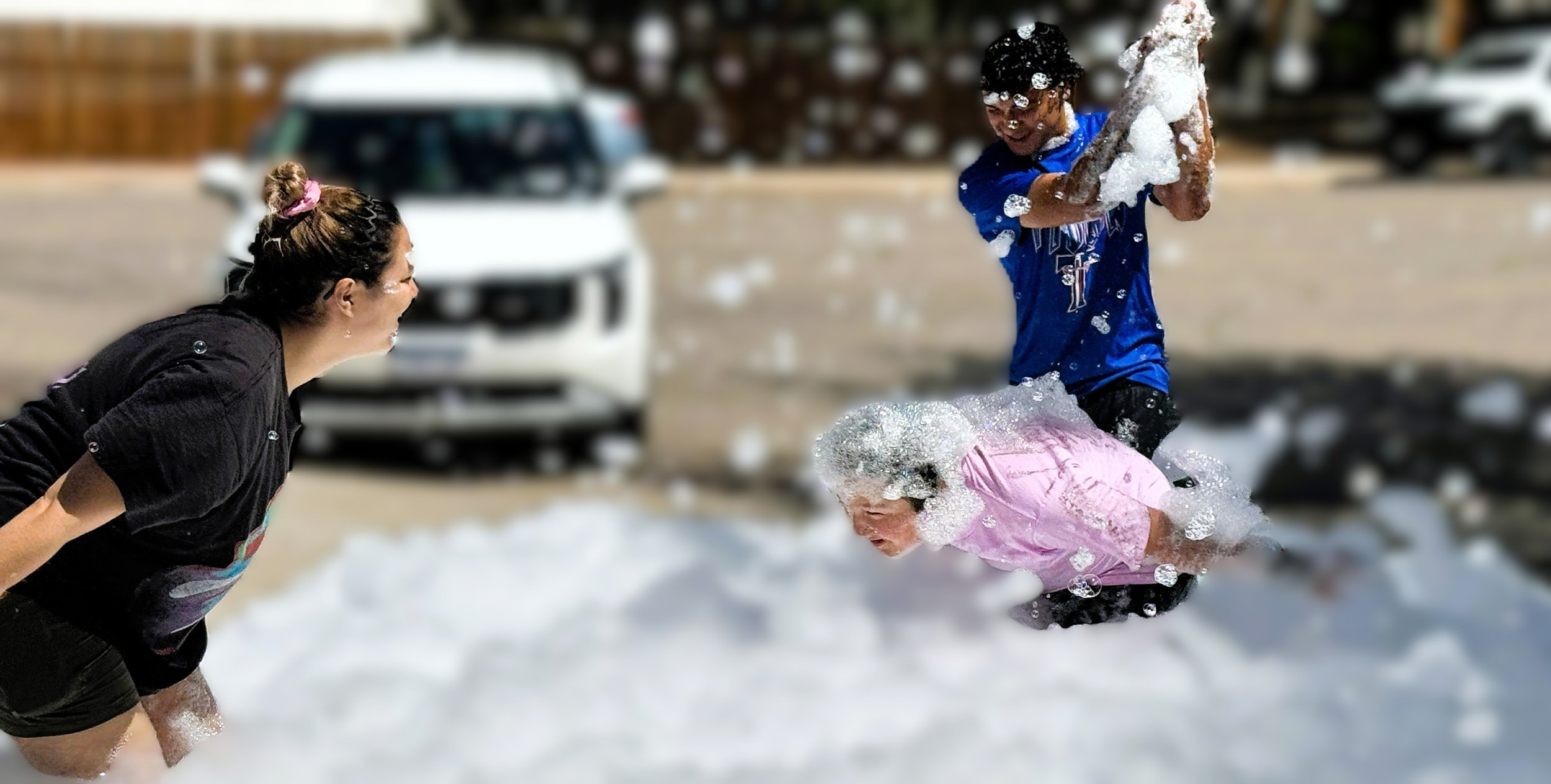 Two children playing in a snowy outdoor area with a woman, likely their mother, laughing and engaging with them. One child is jumping into the snow, and the other is bending down, all enjoying winter activities.
