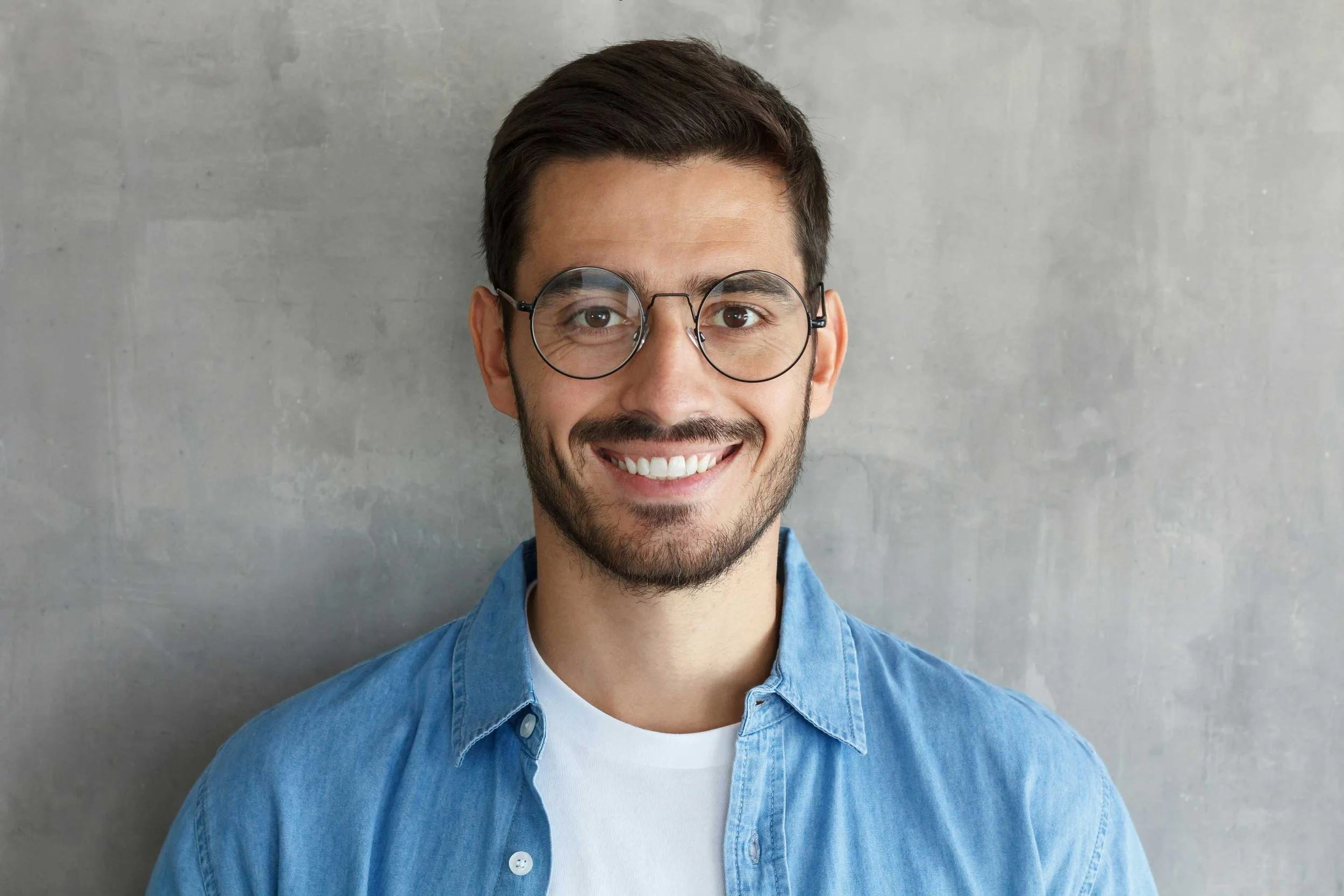 Ein junger Mann mit braunen Haaren, Brille und einem freundlichen Lächeln, trägt ein blaues Hemd und steht vor einer grauen, feinkörnigen Wand.