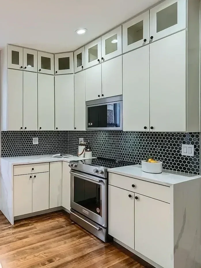 Modern kitchen with white cabinets, black knobs, black hexagon tile backsplash, stainless steel stove, microwave, and wooden floor.