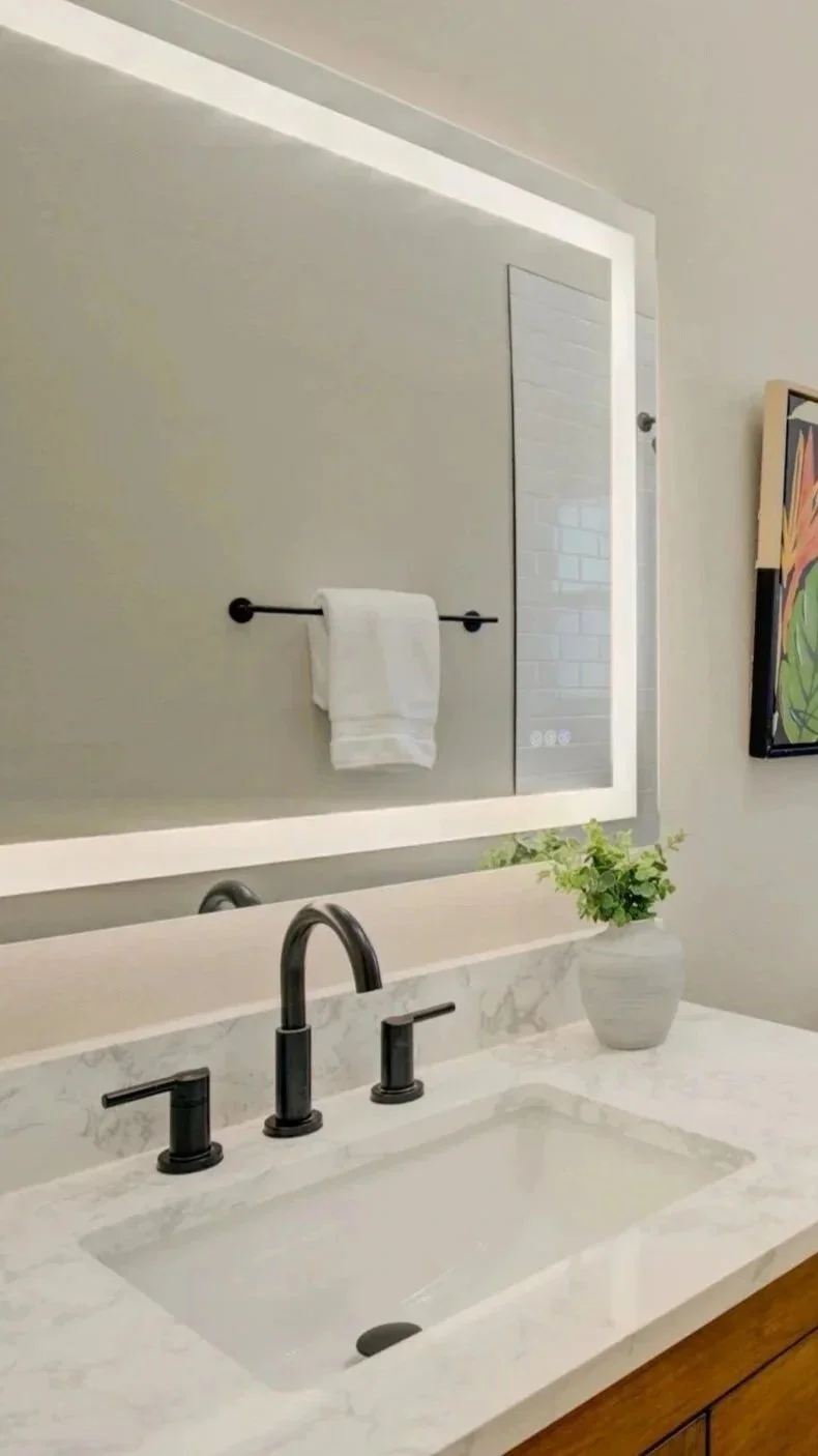 Modern bathroom sink with black faucet and handles, white marble countertop, and a potted plant. A mirror with LED lighting, a towel rack with a white towel, and a colorful wall art are visible in the background.
